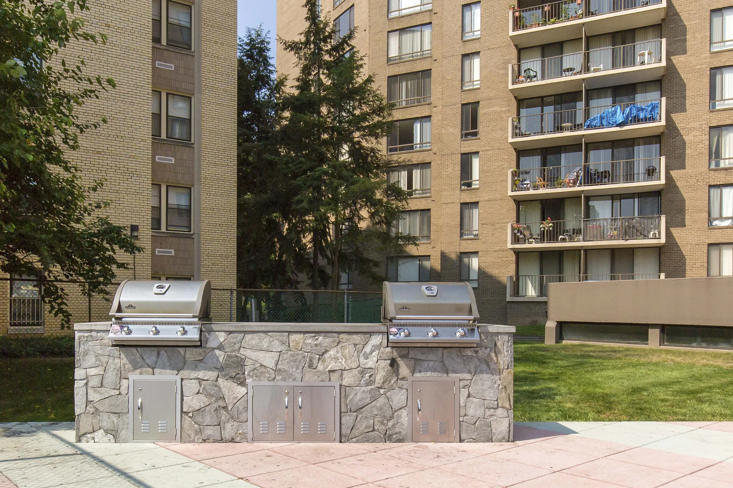 Inviting outdoor patio area with modern barbecue grills and comfortable sun lounger, set against a backdrop of lush trees and stylish apartment buildings.