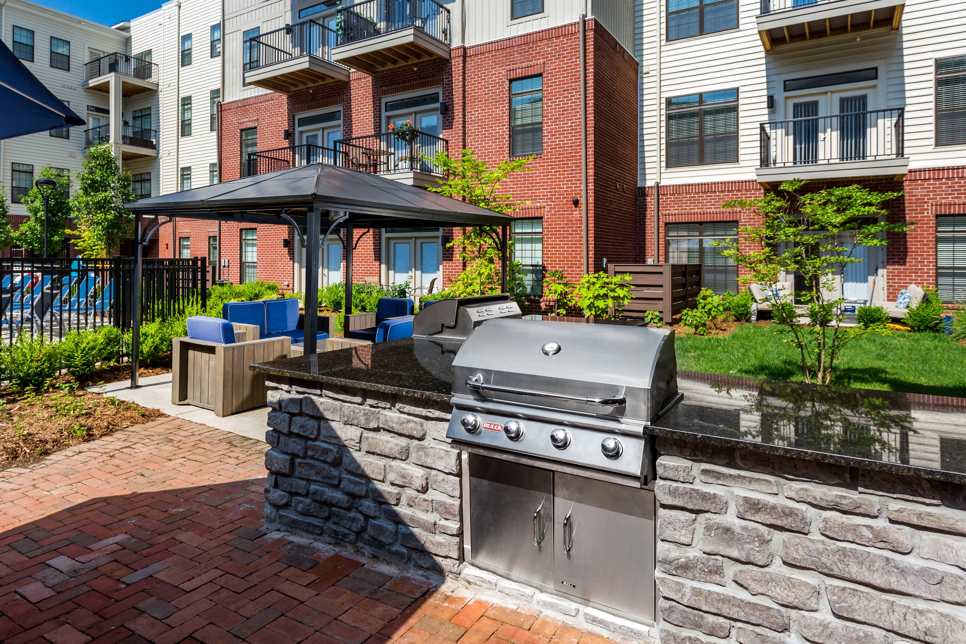 Chic outdoor space featuring a modern stainless steel grill, surrounded by lush greenery and stylish patio seating, set against elegant red brick apartment exteriors.