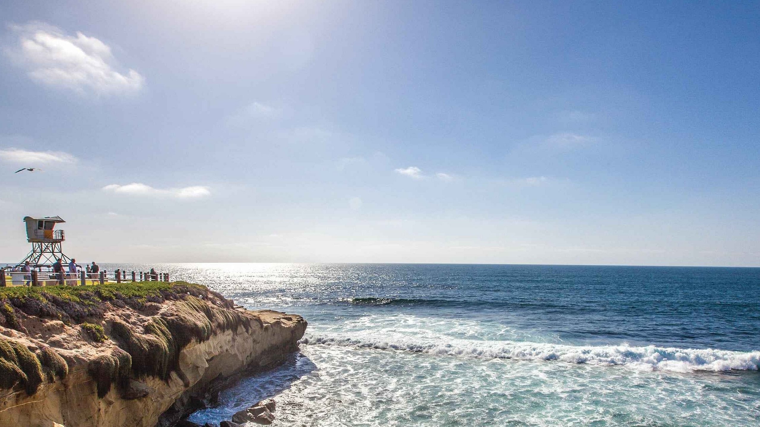 Scenic coastal view showcasing vibrant greenery, dramatic cliffs, and a serene ocean under a clear blue sky with a watchtower in the distance.