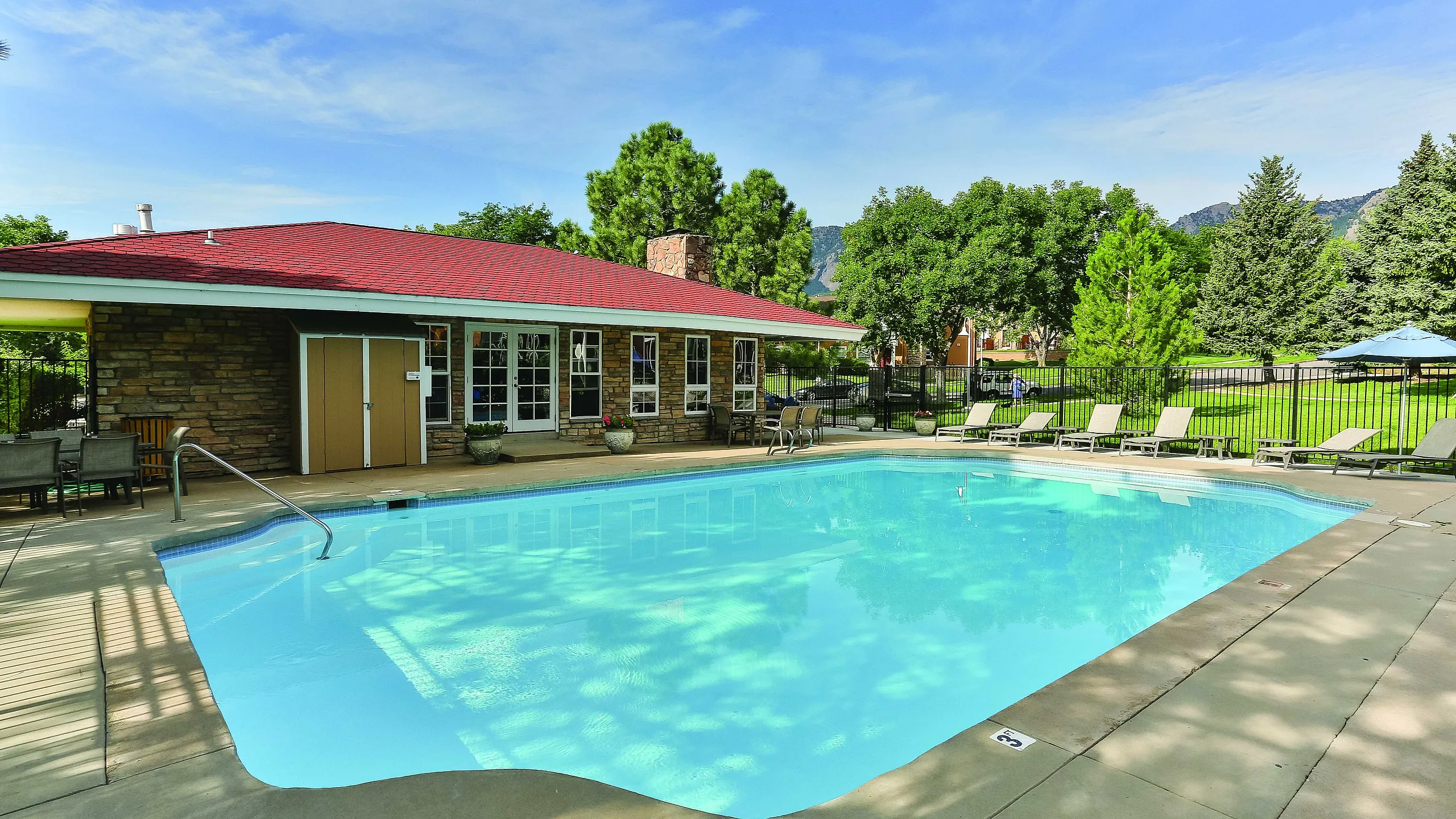 Inviting outdoor pool area with crystal-clear water, surrounded by comfortable lounge chairs and lush greenery, adjacent to a charming brick clubhouse with a red roof.