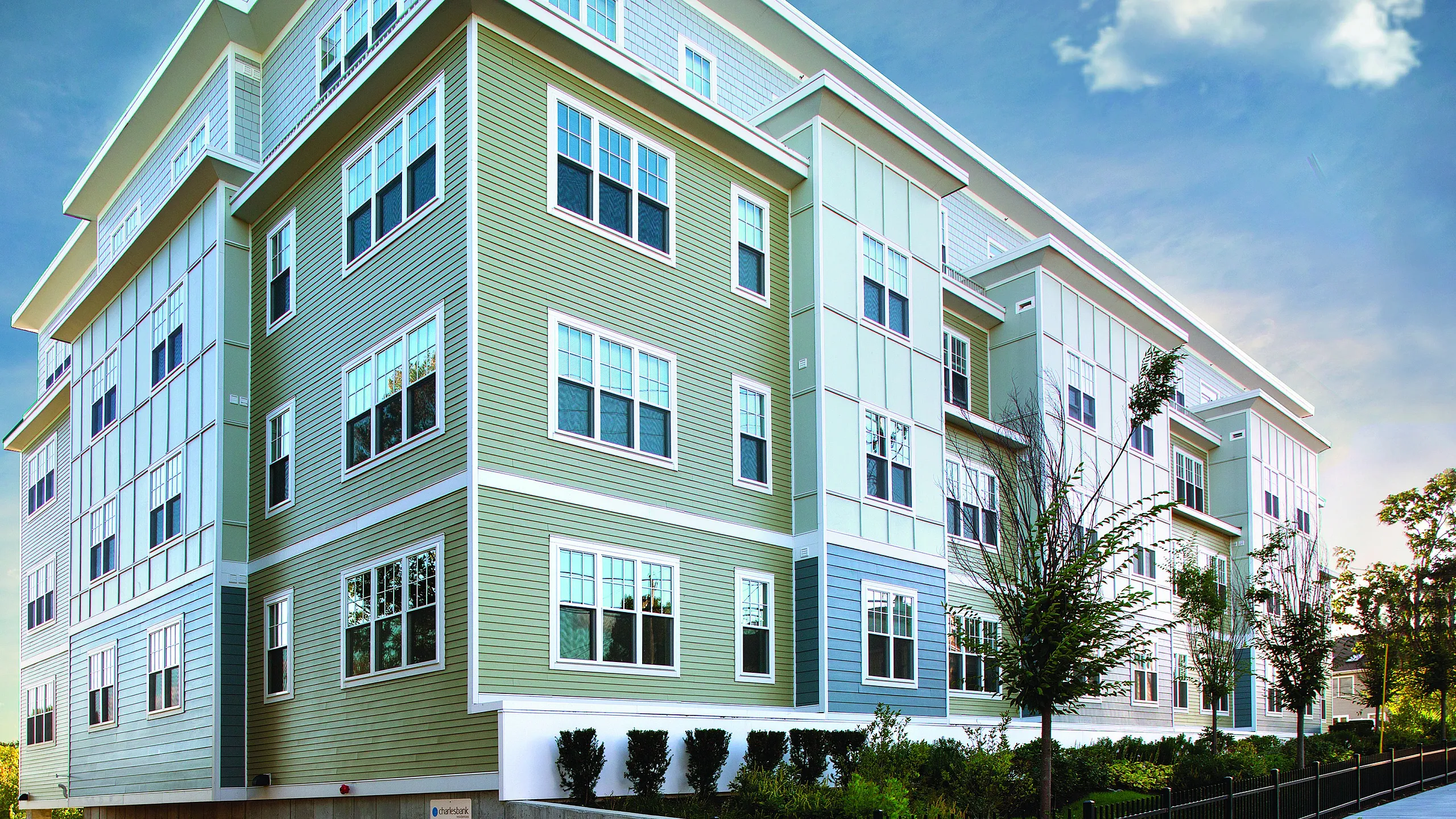 Modern apartment building with stylish green and white exterior, large windows, and lush landscaping under a clear blue sky.