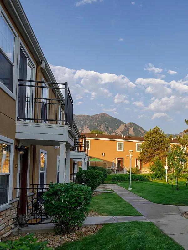Charming apartment complex with scenic pathways, lush greenery, and stunning mountain views under a clear sky.