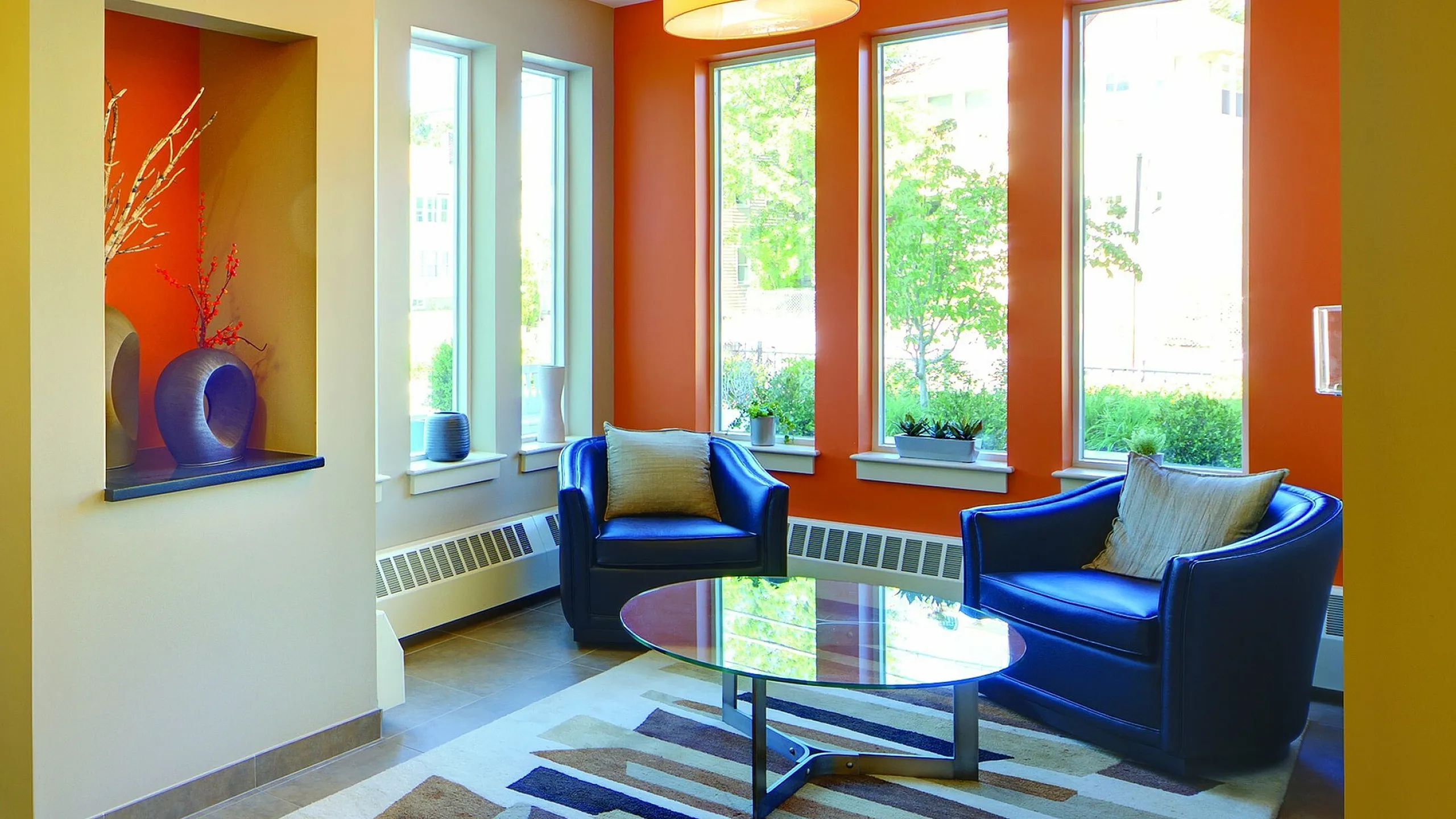 Inviting apartment lobby with modern decor, vibrant orange accent wall, and comfortable seating area featuring blue armchairs and a stylish glass coffee table.