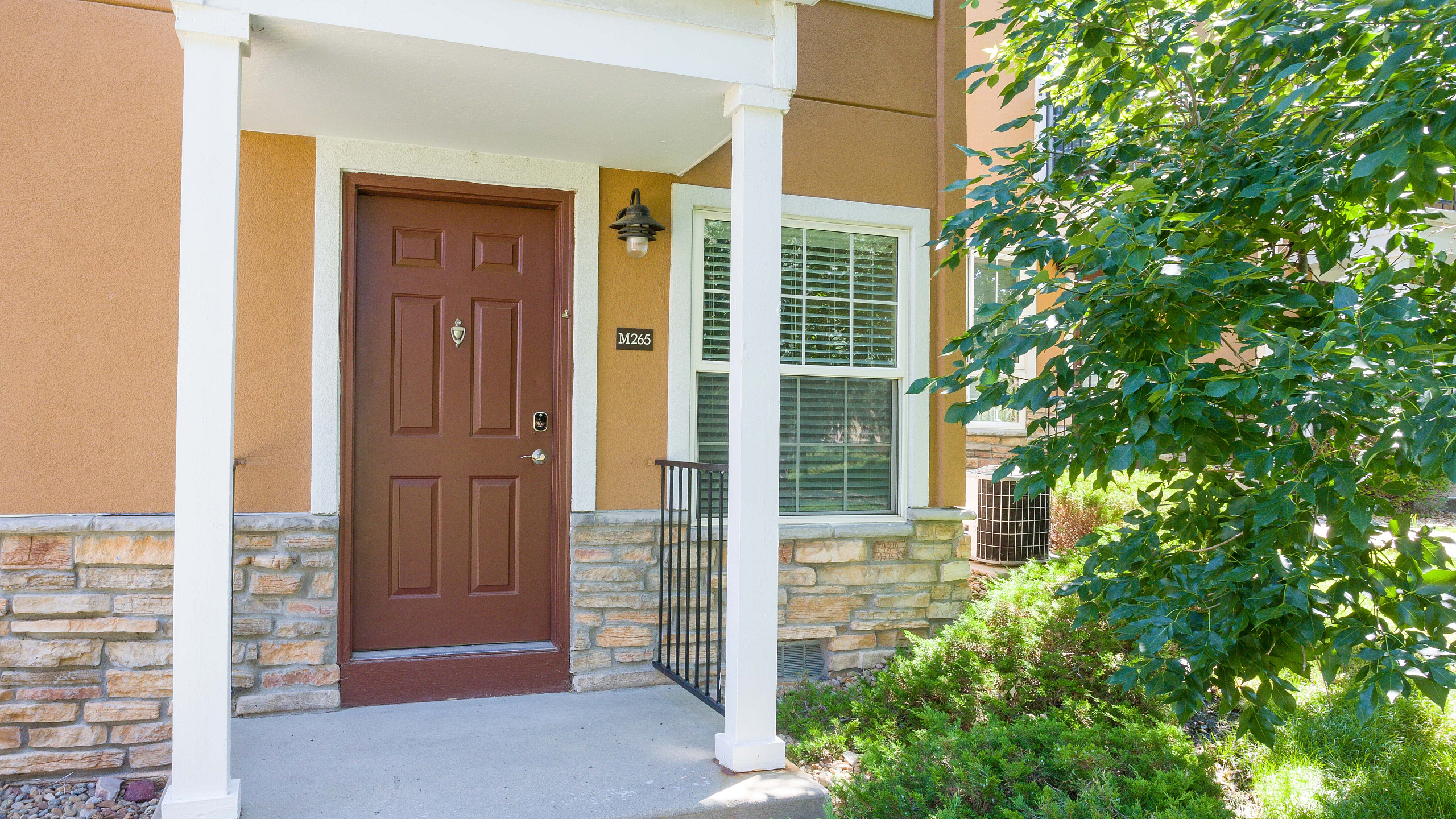 Inviting apartment entrance with a warm brown door, framed by elegant white pillars and surrounded by lush greenery.