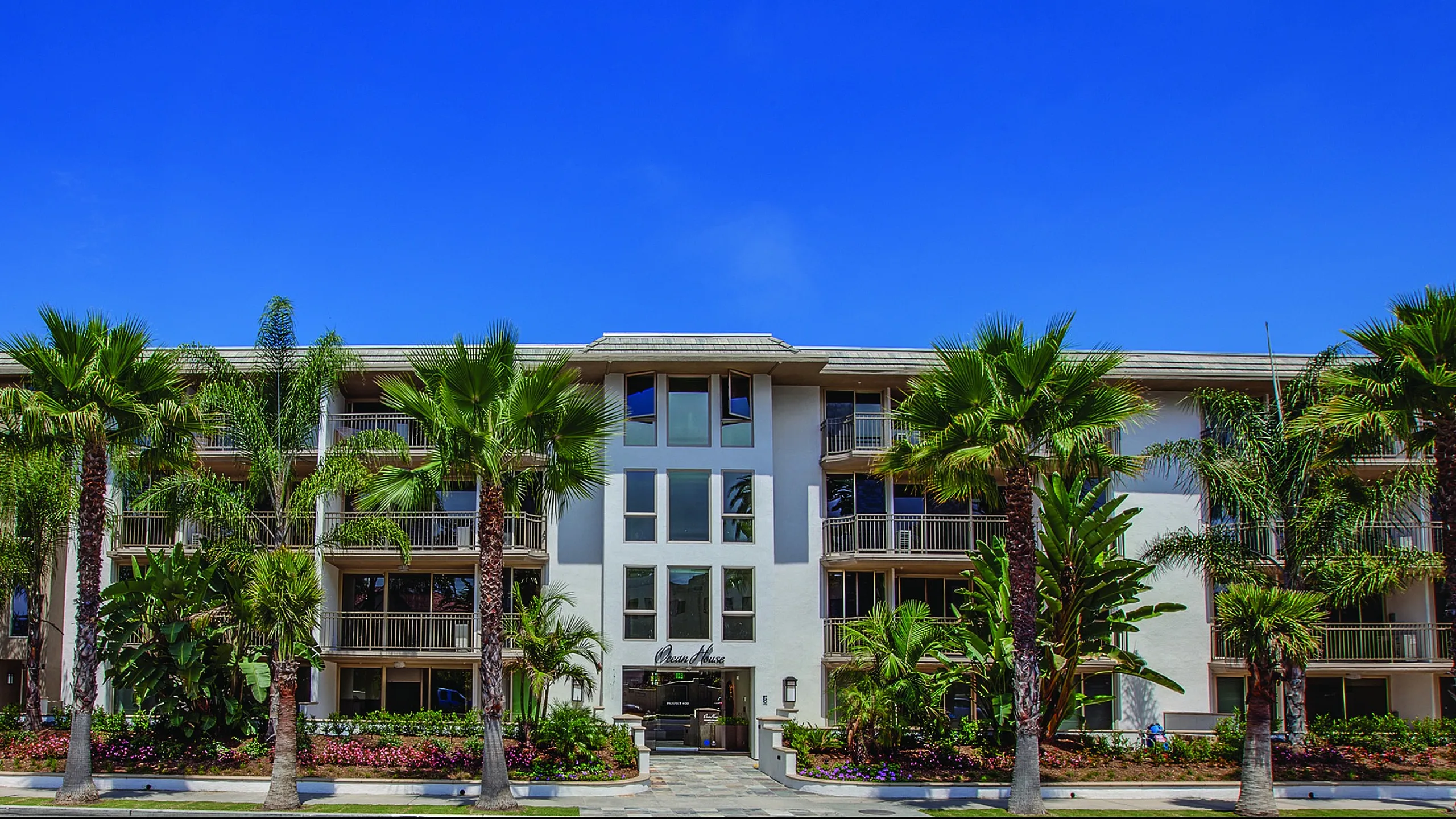 Elegant apartment building with lush palm trees, stunning architecture, and vibrant landscaping under a clear blue sky.