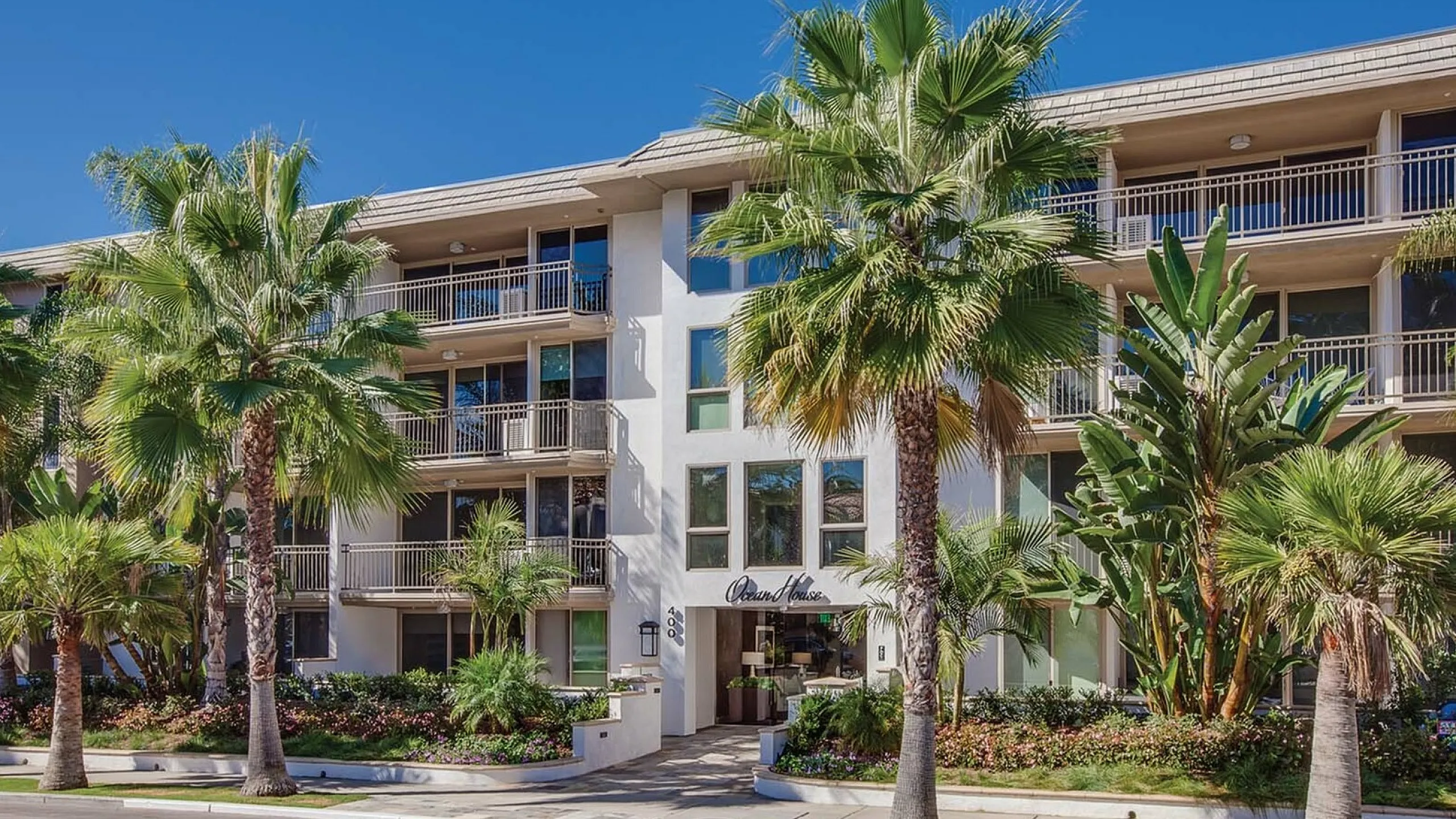 Beautiful apartment building with elegant balconies surrounded by lush palm trees and vibrant landscaping under a clear blue sky.