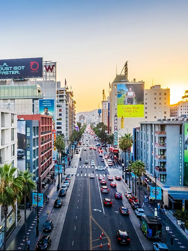 Vibrant cityscape view showcasing modern apartments with artistic billboards, palm-lined streets, and bustling urban life at sunset.