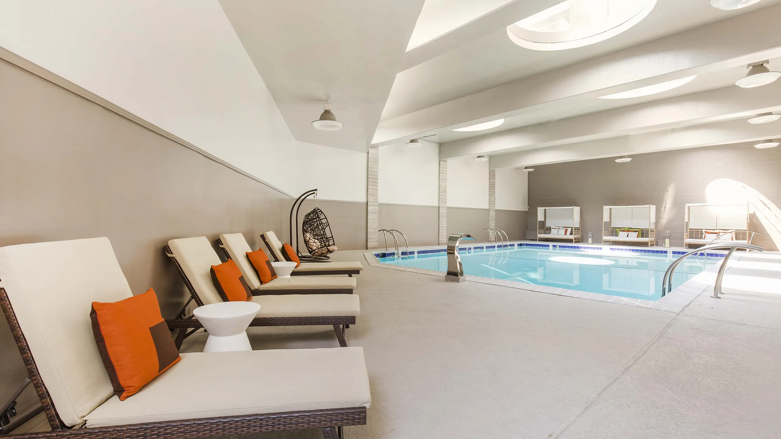 Luxurious indoor swimming pool area with elegant lounge chairs, vibrant orange accents, and skylight design for natural illumination.