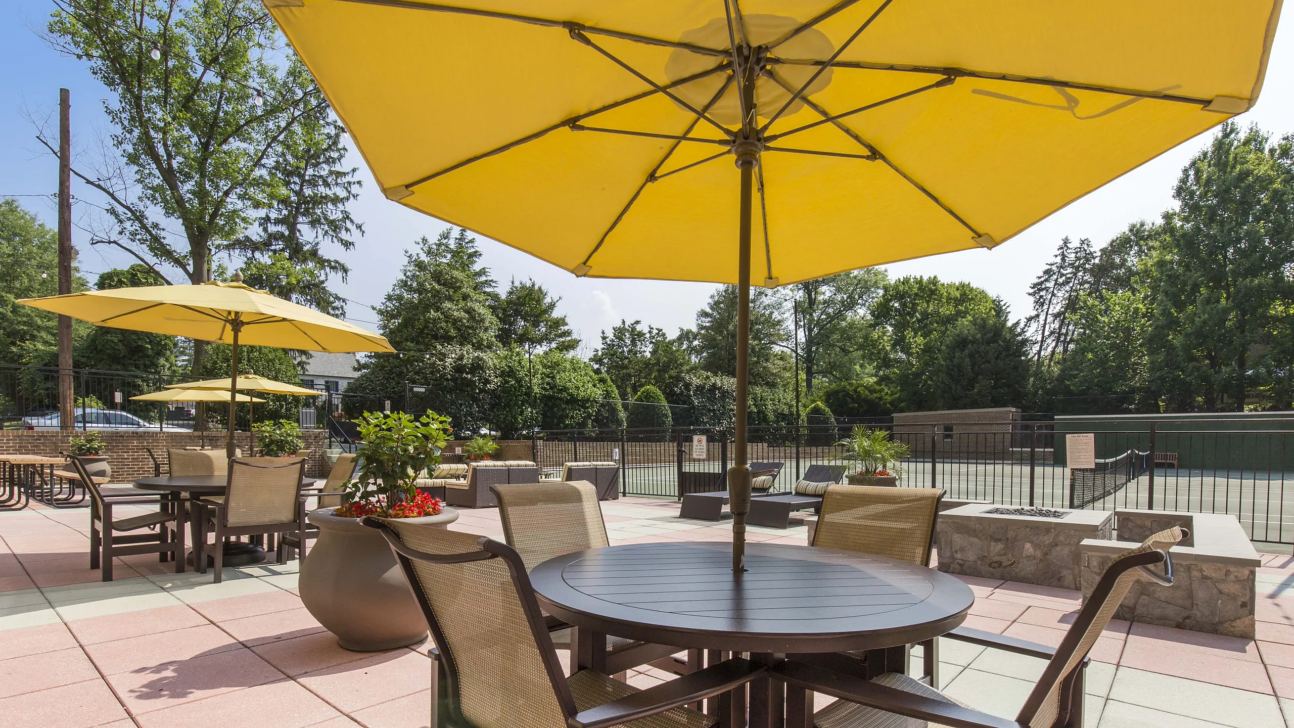 Vibrant outdoor patio with stylish seating under yellow umbrellas, lush greenery, and a tennis court backdrop.