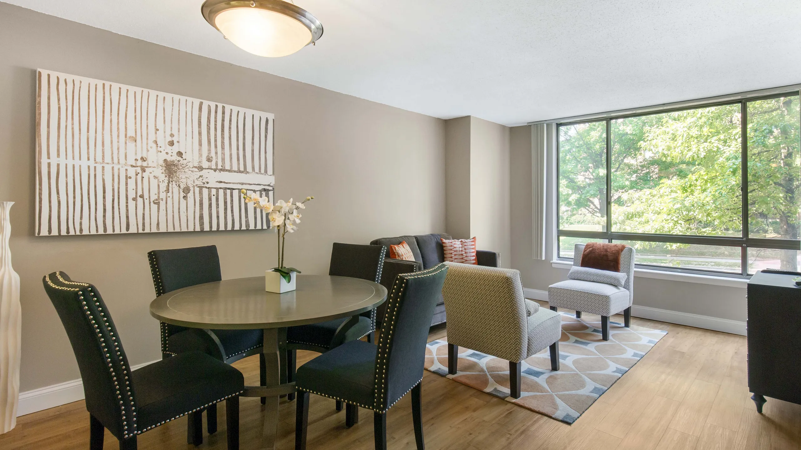 Bright and inviting apartment living area with stylish decor, featuring a cozy seating arrangement and a dining area under elegant lighting, set against large sunlit windows and warm wooden flooring.