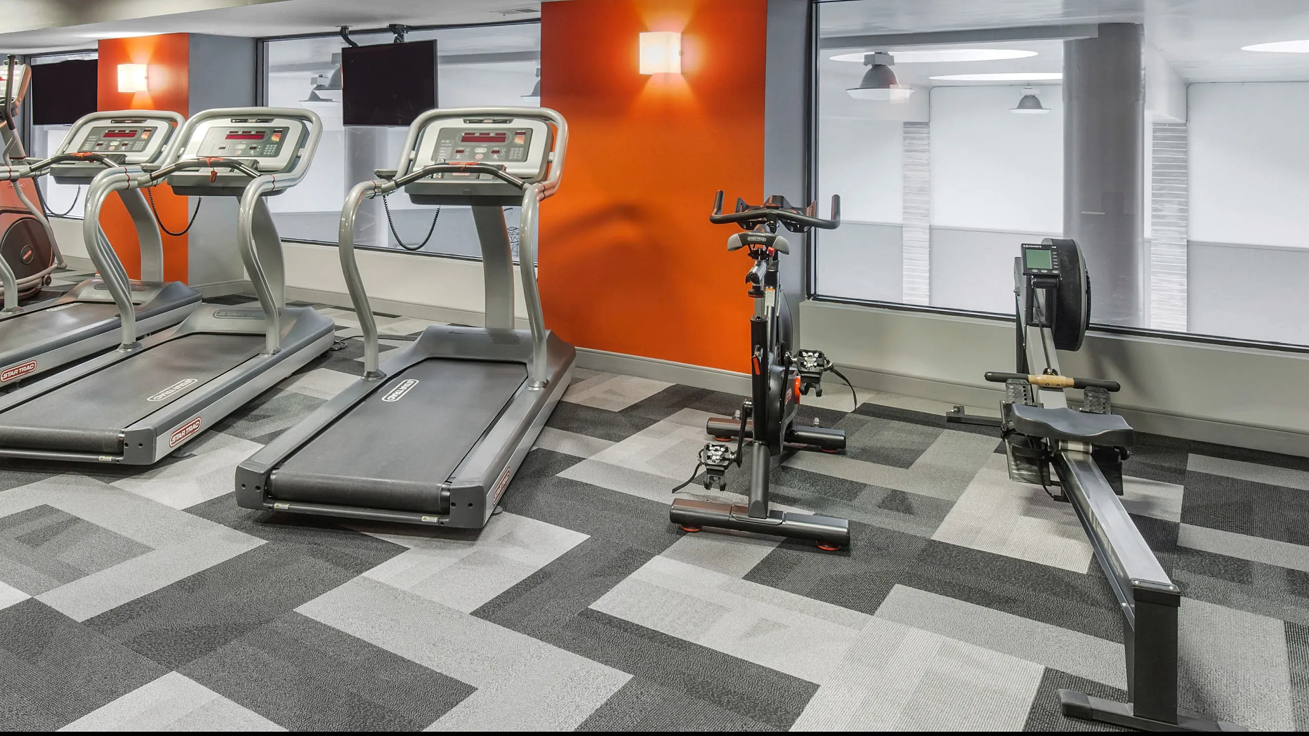 Modern gym with sleek treadmills, exercise bike, and rowing machine against vibrant orange accents and large windows.