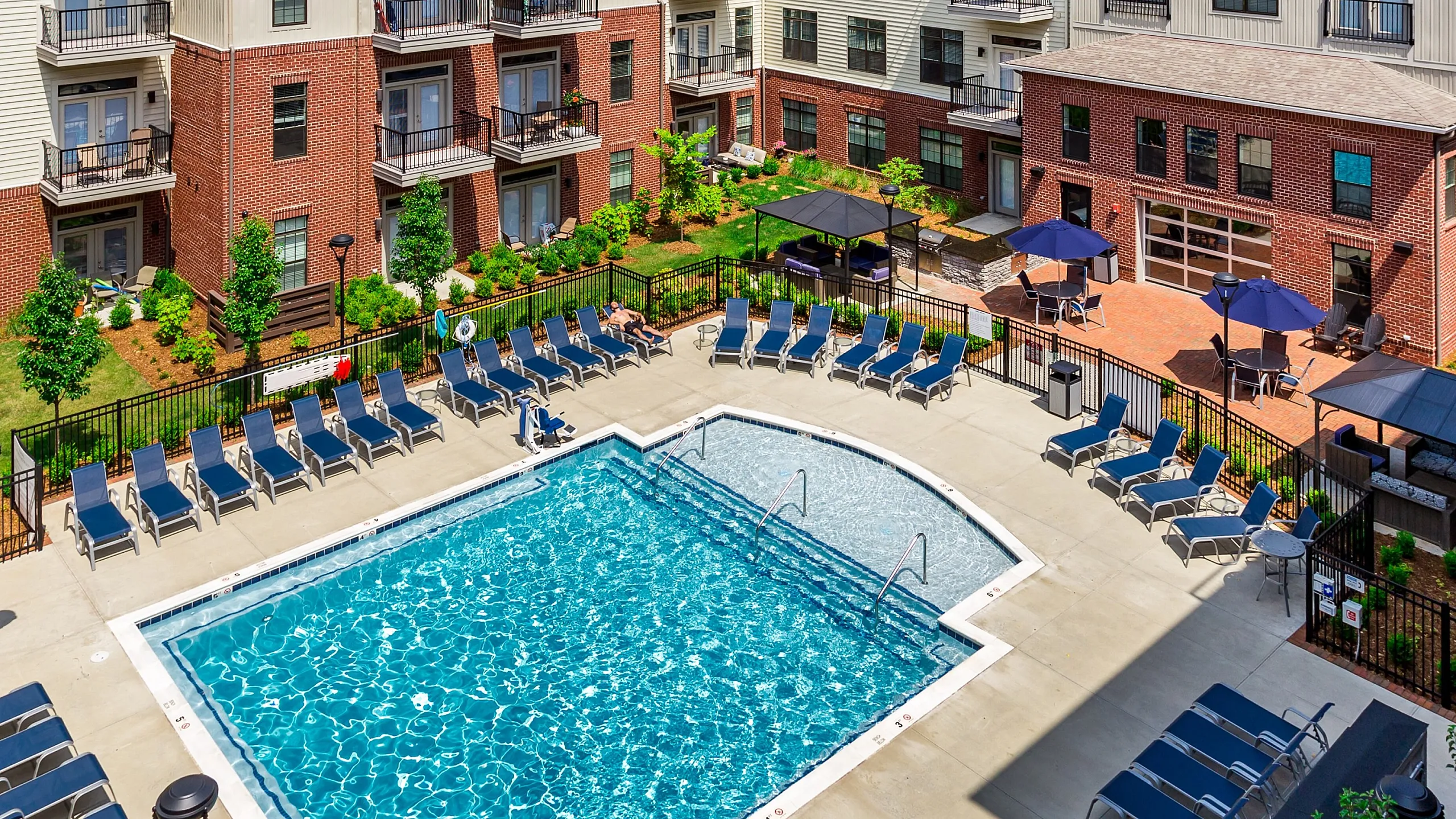 Luxurious apartment complex featuring a stunning outdoor pool with ample lounge seating, surrounded by elegant multi-story buildings and vibrant landscaped gardens.