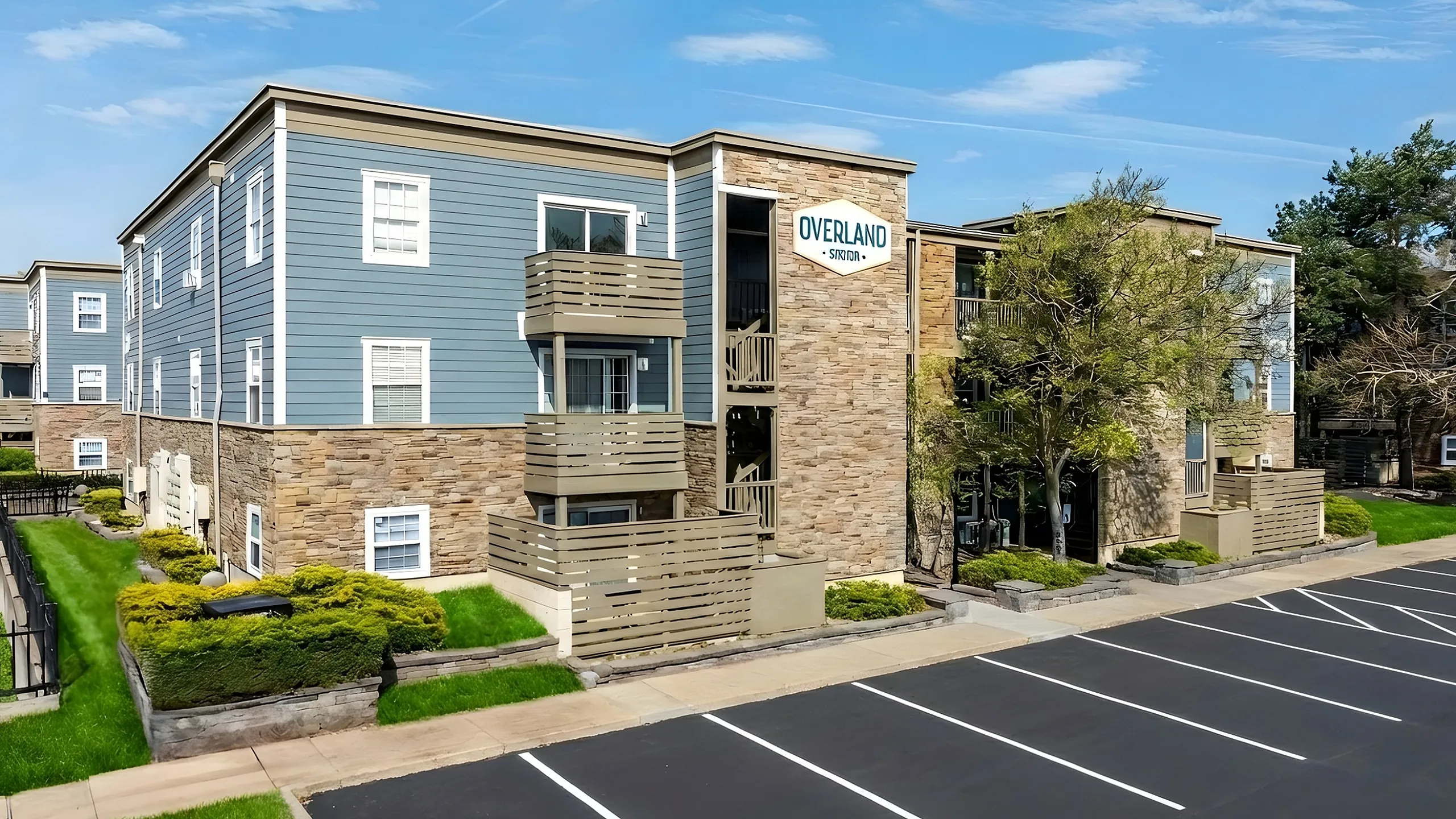 Modern apartment complex with stylish blue and stone facade, featuring private balconies, lush greenery, and ample parking spaces.