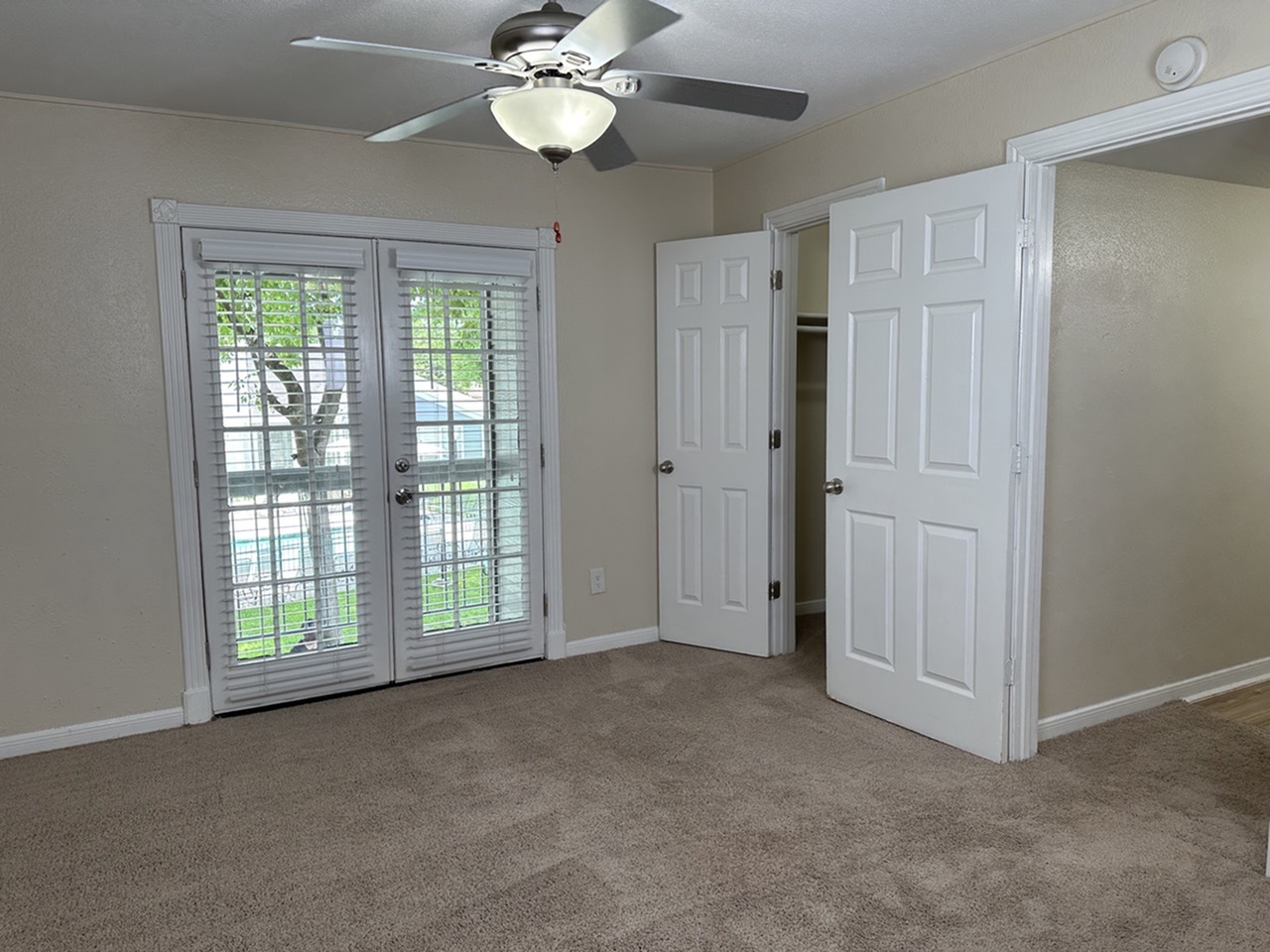 Bright bedroom with plush carpet, French doors, and a ceiling fan, offering comfort and style.