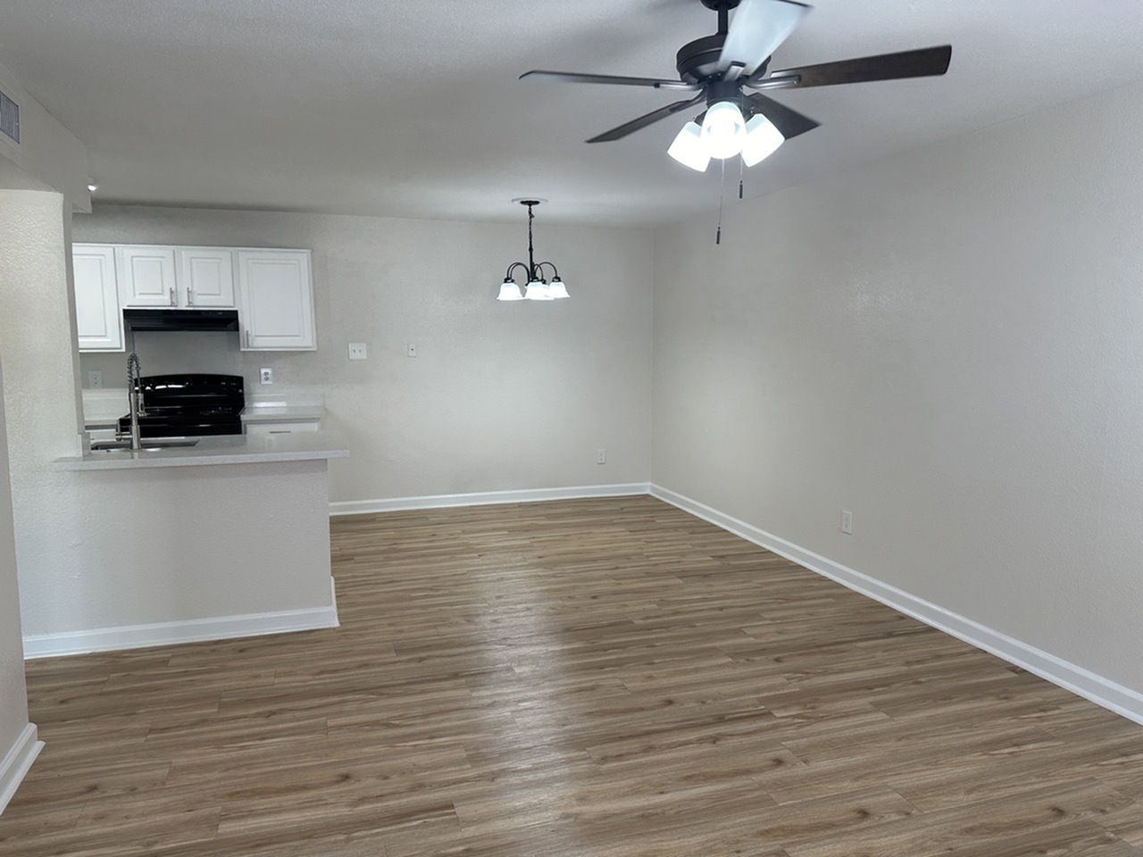 Modern open-plan living area with sleek wood-style floors, elegant ceiling fan, and a compact kitchen featuring white cabinets and black appliances.
