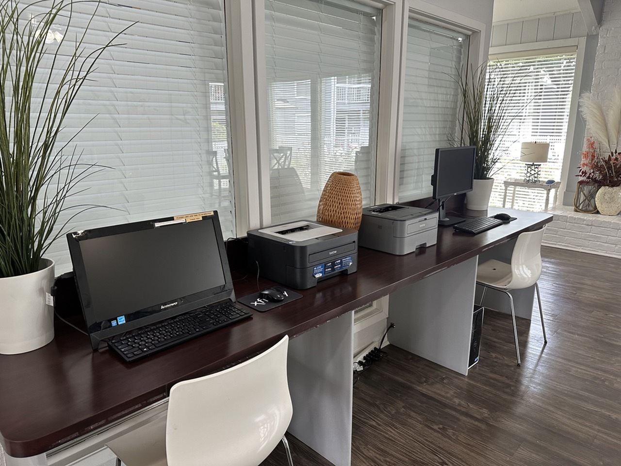 Modern communal workspace featuring sleek computers and printers on a polished wooden desk, complemented by natural light and stylish greenery.