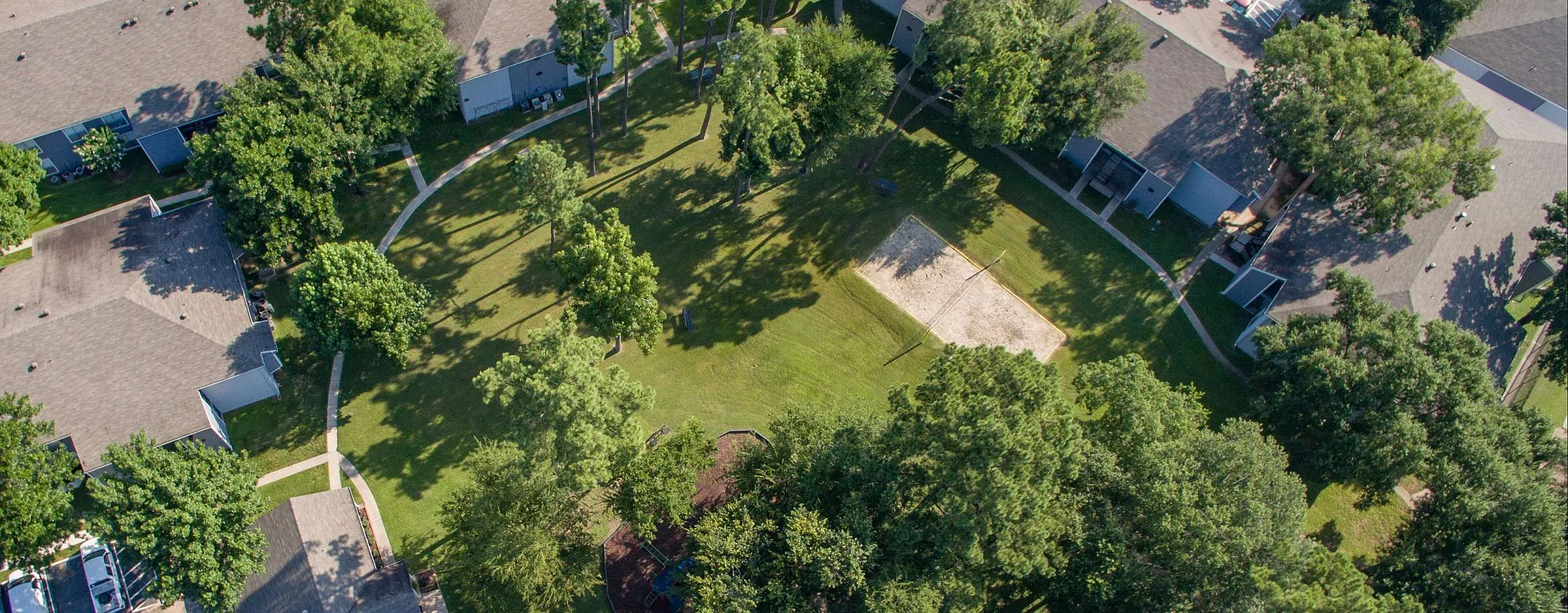 Aerial view of a serene apartment complex surrounded by lush greenery, featuring a central park with a playground and ample parking space.