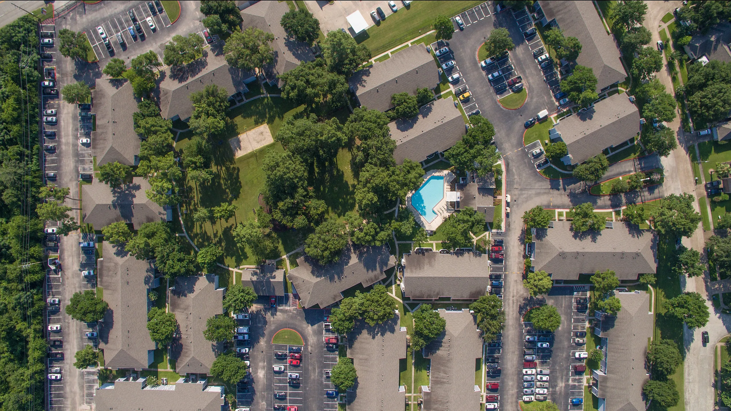 Aerial view of a lush apartment complex featuring spacious lawns, a sparkling pool, and ample parking facilities amidst tree-lined pathways.