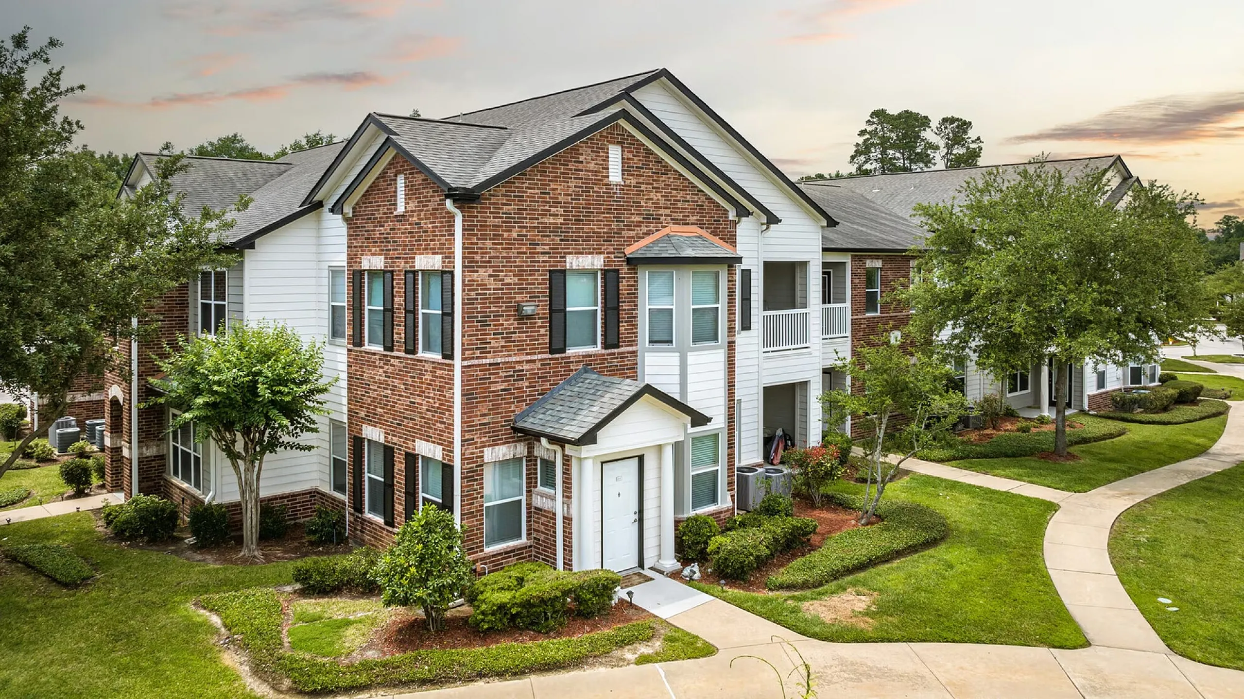 Charming red brick apartment building with manicured lawns and a serene walkway under a colorful sunset sky.