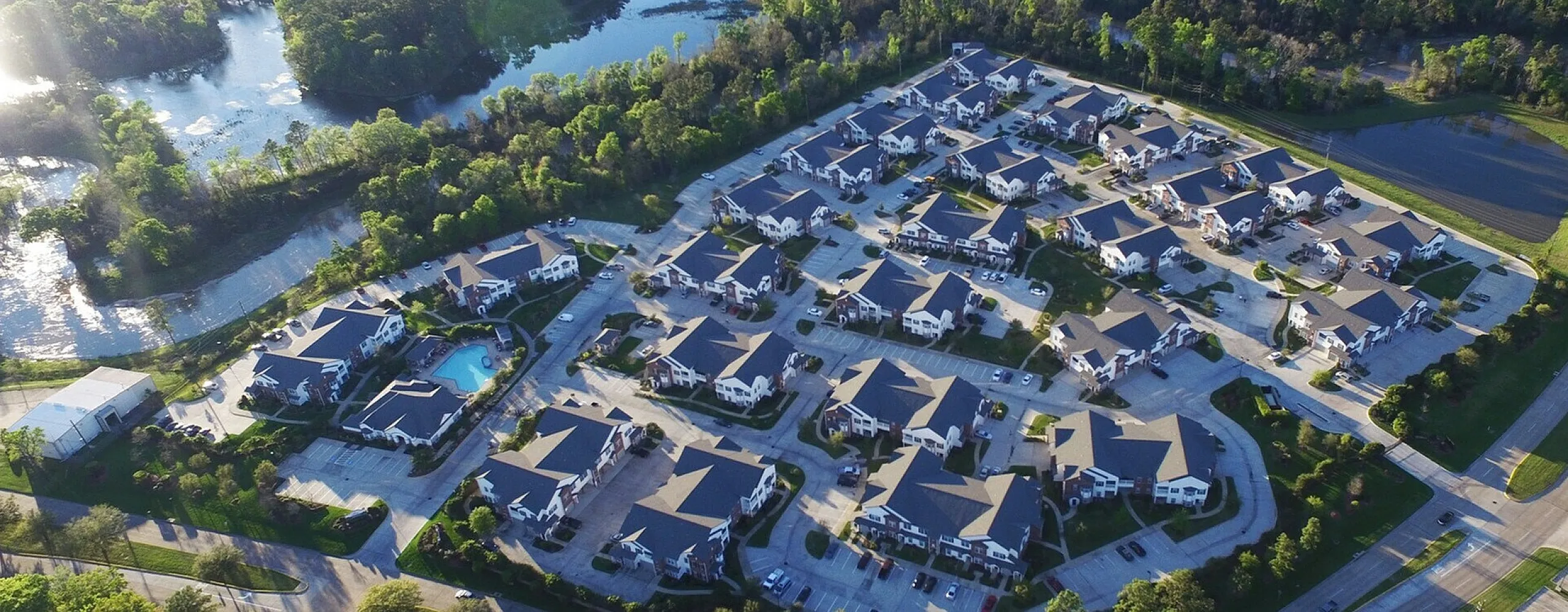Aerial view of a picturesque apartment community nestled in lush greenery with a serene lake, featuring modern buildings, well-maintained roads, and a sparkling pool.