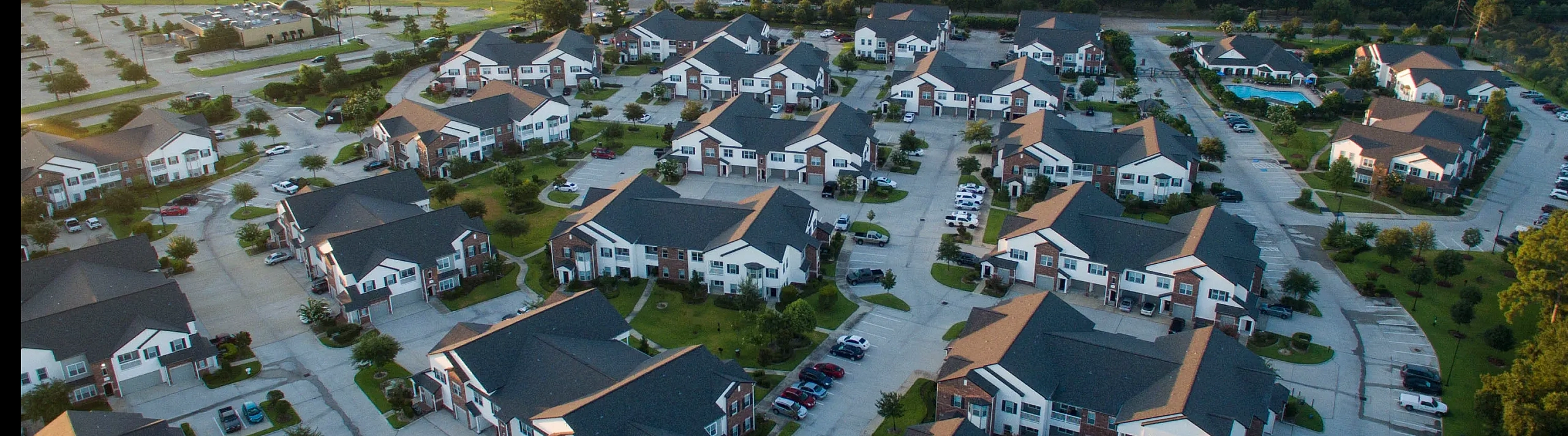 Aerial view of a picturesque apartment community nestled in lush greenery with a serene lake, featuring modern buildings, well-maintained roads, and a sparkling pool.