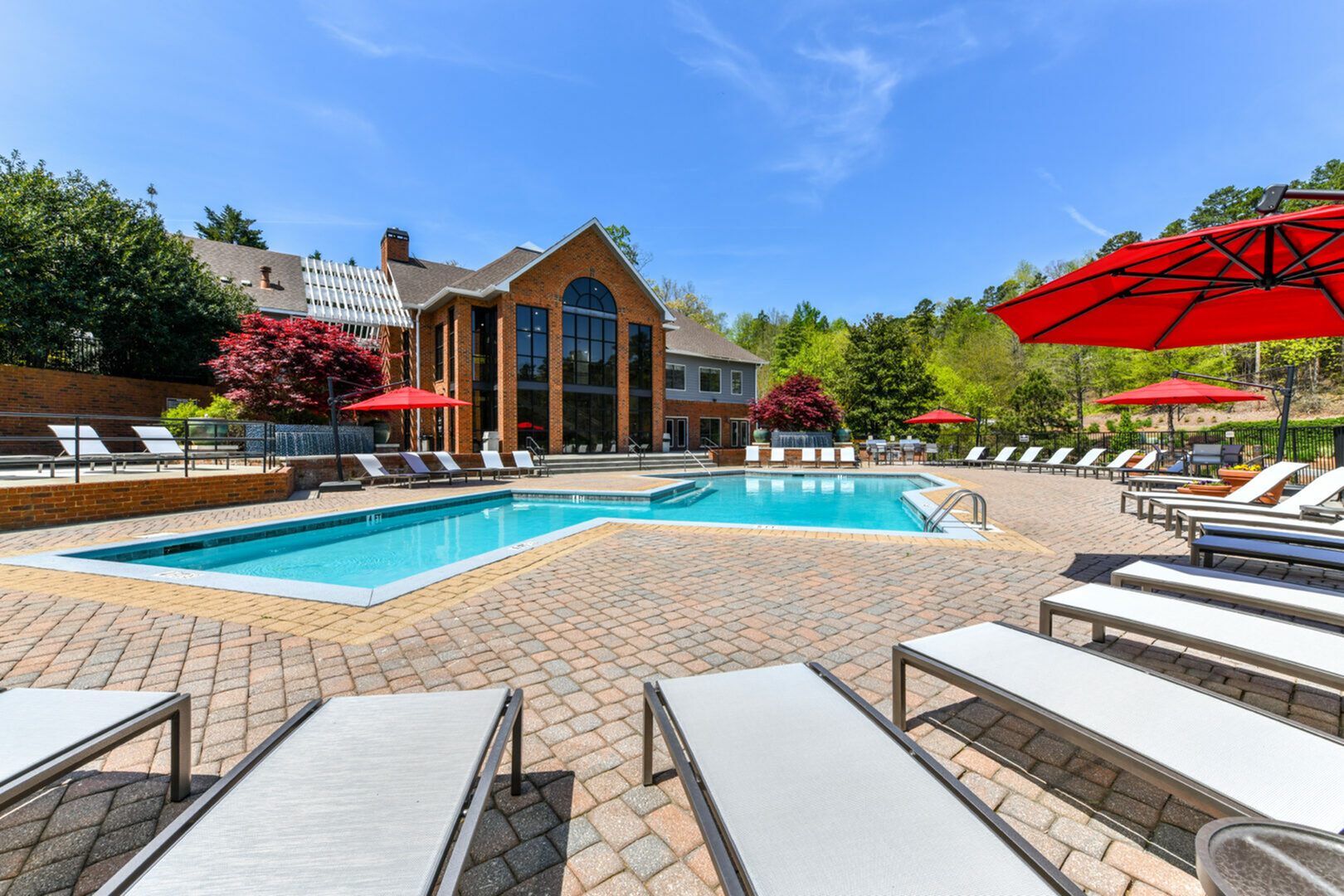 Inviting pool area with cozy lounge chairs, vibrant red umbrellas, and charming brick clubhouse surrounded by lush greenery under a clear blue sky.