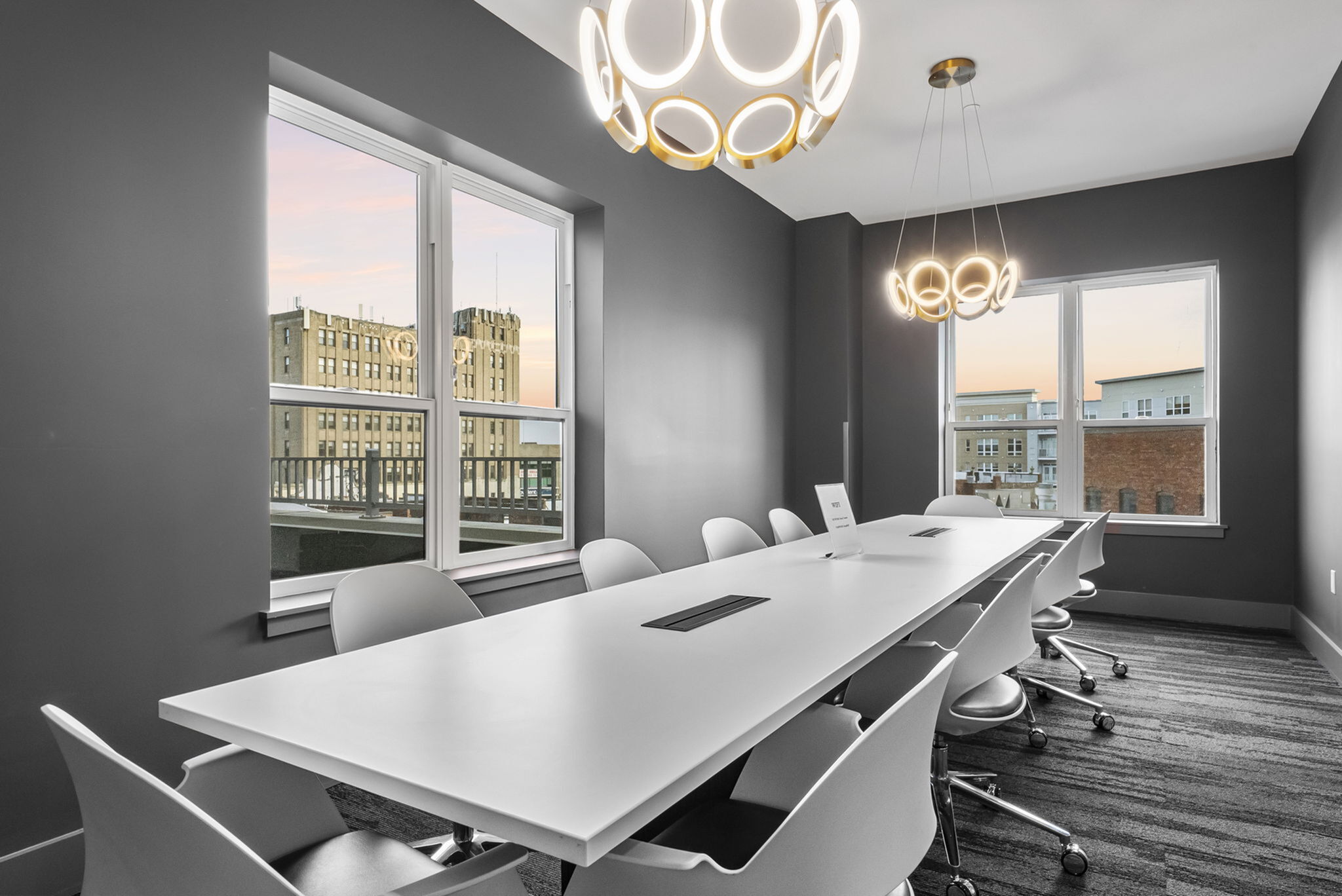 Modern conference room with sleek white table, stylish chairs, and chic pendant lighting, offering cityscape views through large windows.