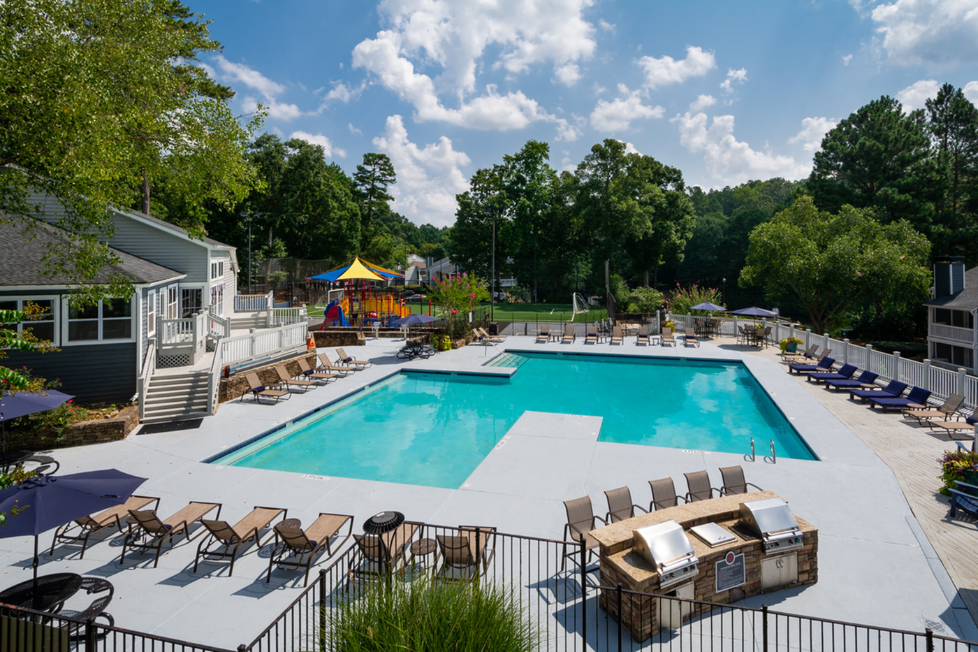 Inviting apartment community showcasing a sparkling swimming pool, luxurious sun loungers, and a vibrant playground surrounded by lush greenery.