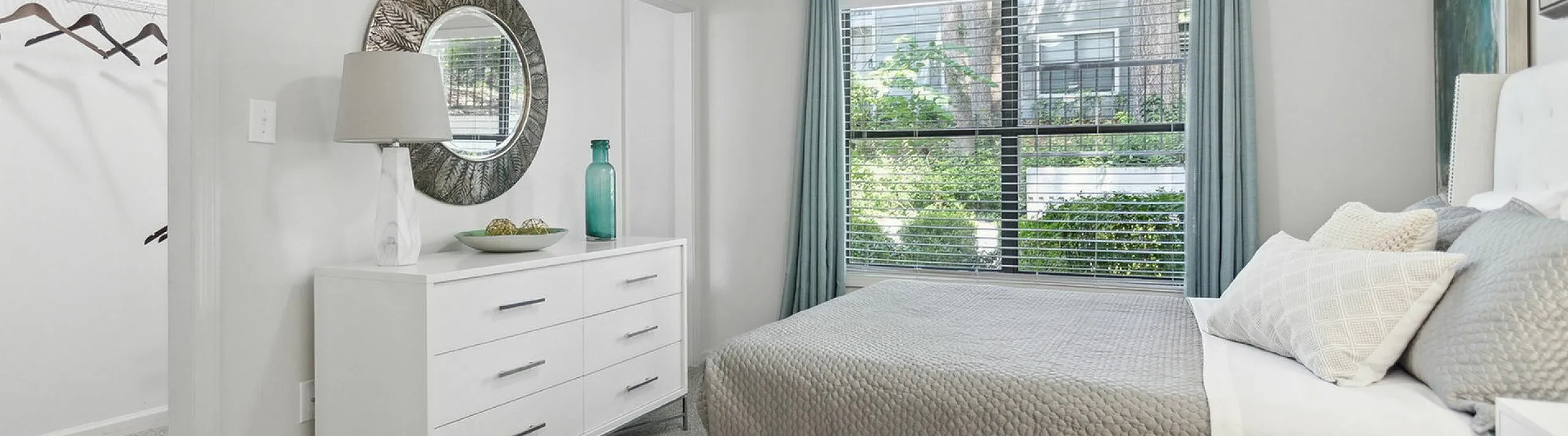 Bright and airy bedroom with a stylish bed, elegant dresser, and large window offering lush green views.
