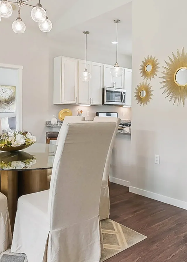 Elegant dining area with stylish glass table, chic upholstered chairs, and modern kitchen in the background with sleek cabinetry and decorative wall mirrors.
