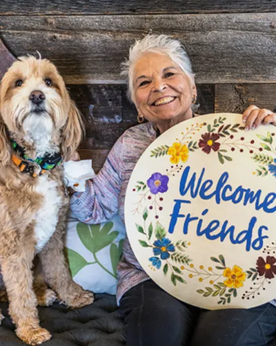 Charming woman holding 'Welcome Friends' sign beside a fluffy dog, radiating warmth and friendliness.