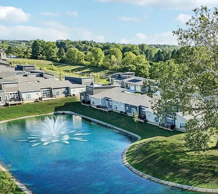 Charming aerial view of modern apartments with scenic pond and fountain, surrounded by lush greenery and open skies.