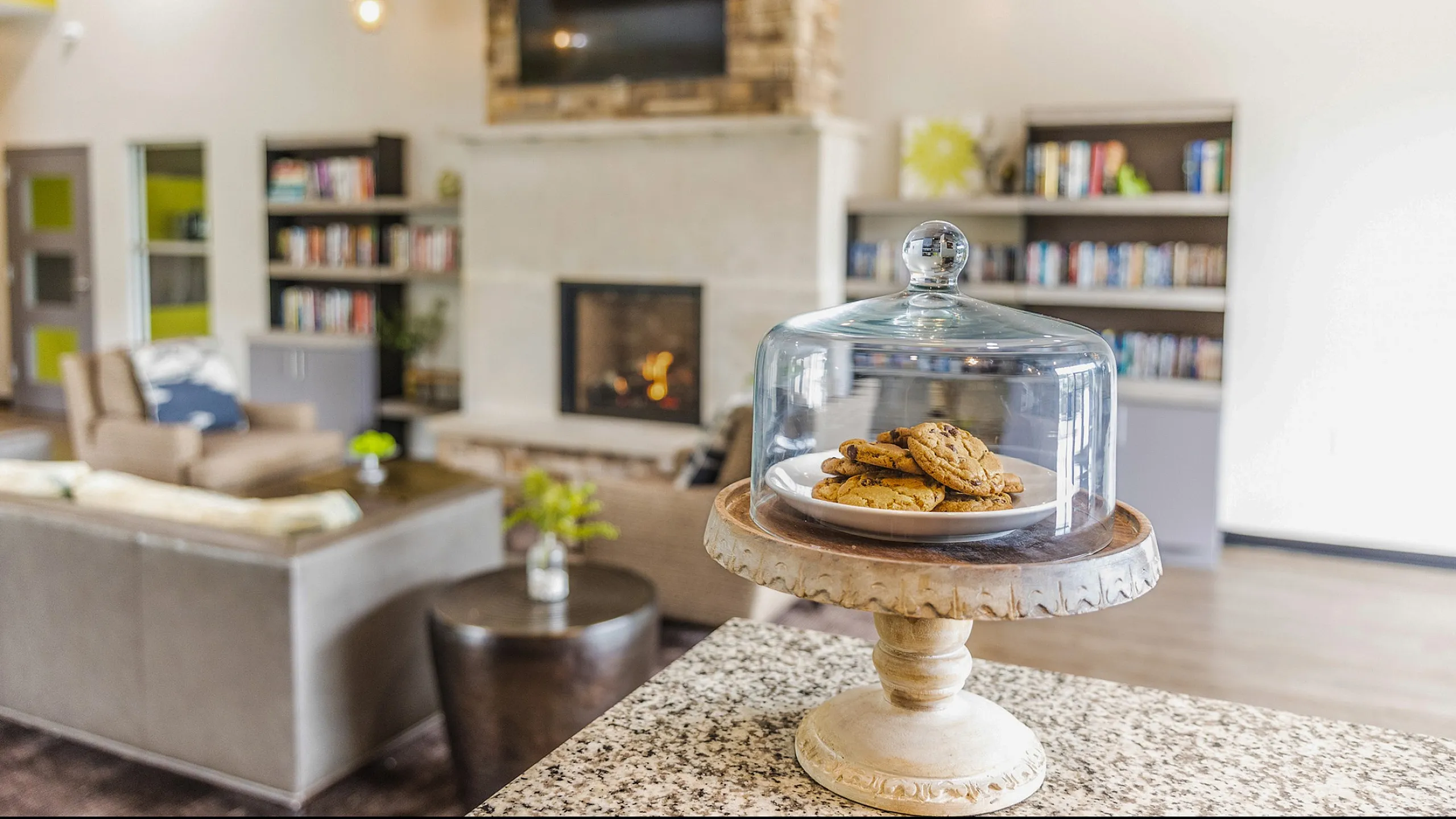 Inviting living room with a cozy fireplace and a modern touch, featuring a stylish bookshelf and freshly baked cookies displayed elegantly under a glass cover.