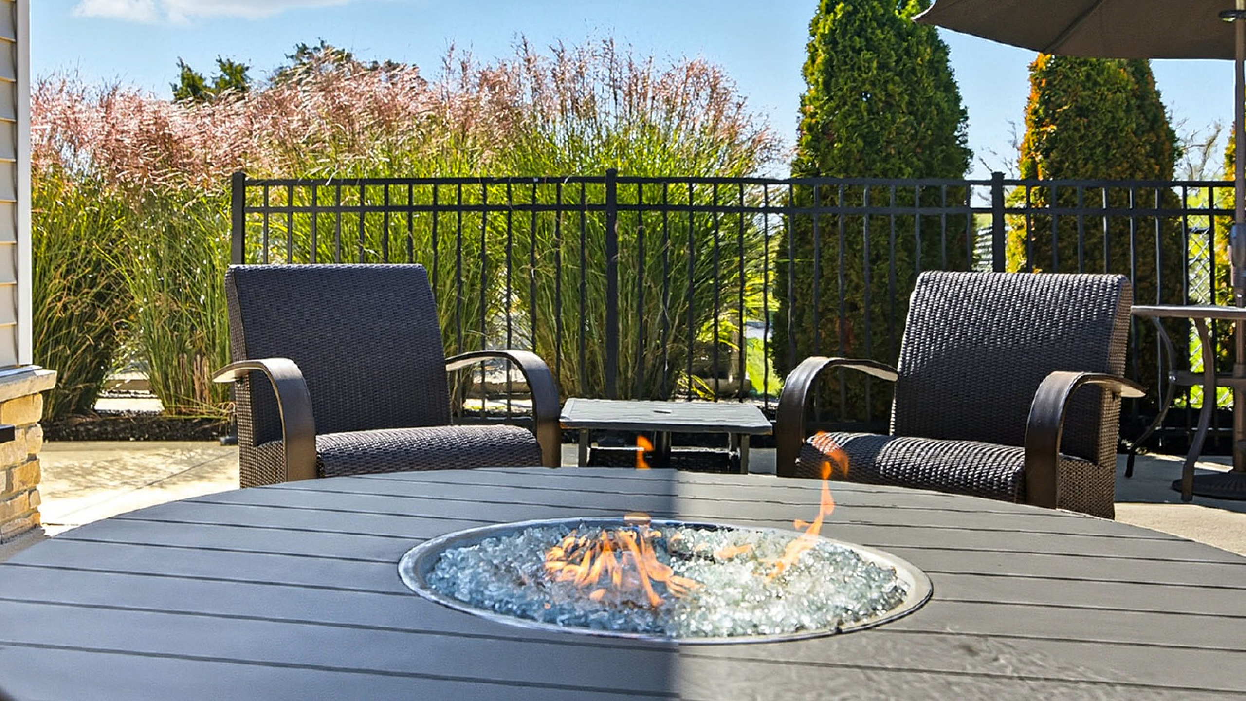 Charming outdoor patio with elegant wicker chairs around a modern fire pit, set against lush greenery and a clear sky.