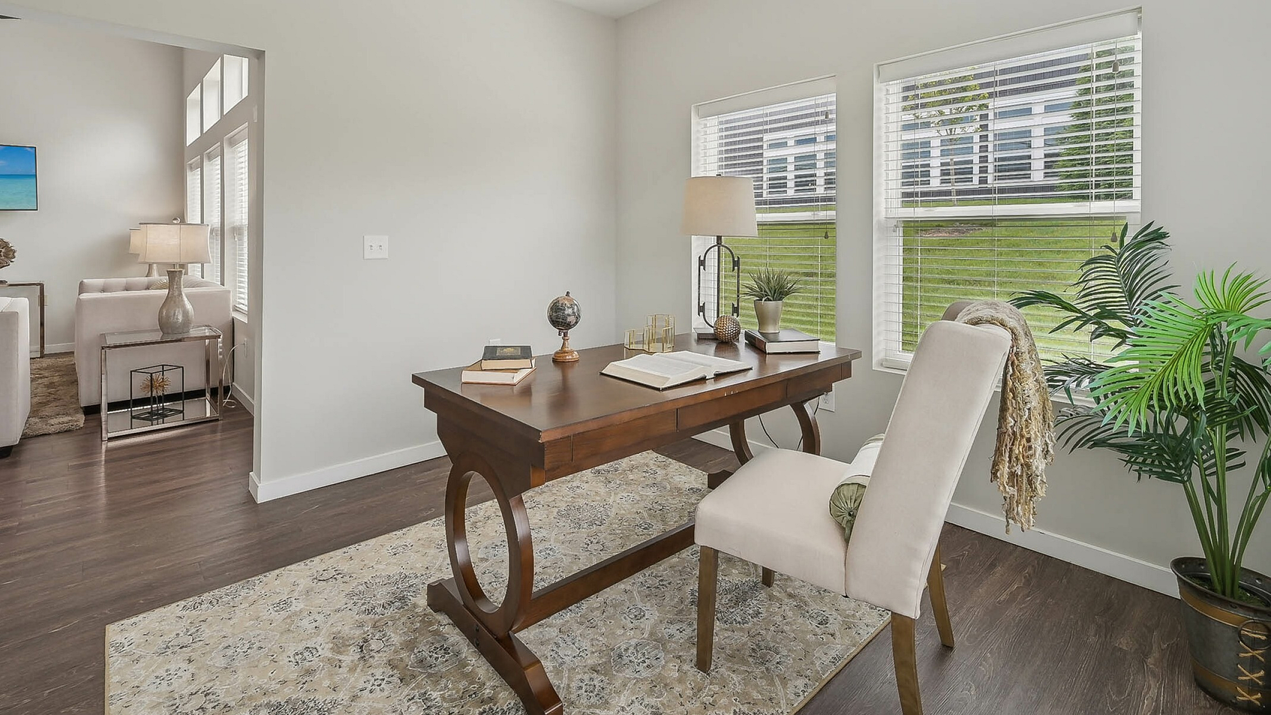 Inviting home office with large windows, elegant wooden desk, and cozy seating area opening into a bright living room with plush sofas.