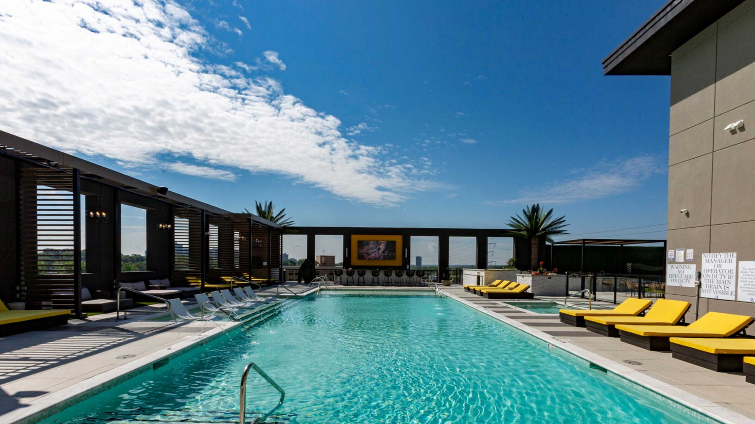 Luxurious rooftop pool with yellow sun loungers, surrounded by modern architecture and vibrant palm trees, under a clear blue sky.