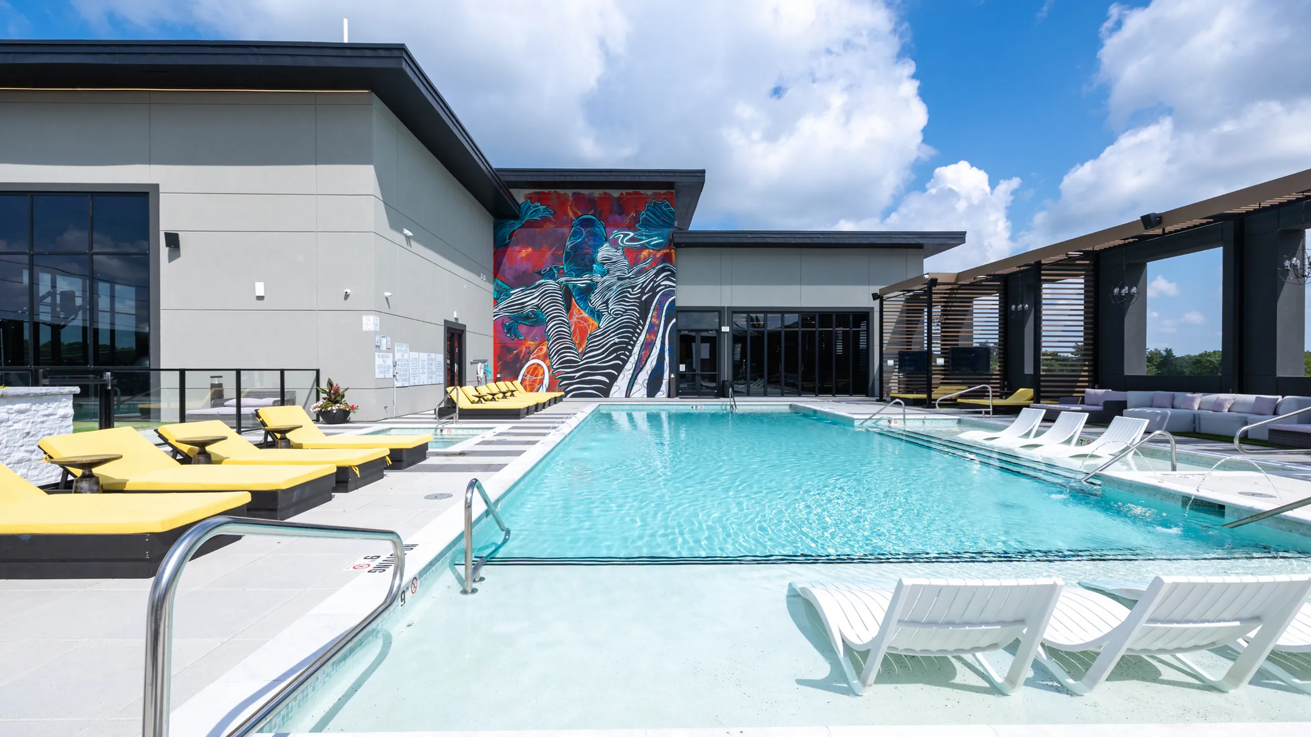 Luxurious modern apartment complex showcasing a stunning pool with vibrant mural, stylish lounge chairs, and a sunny sky.
