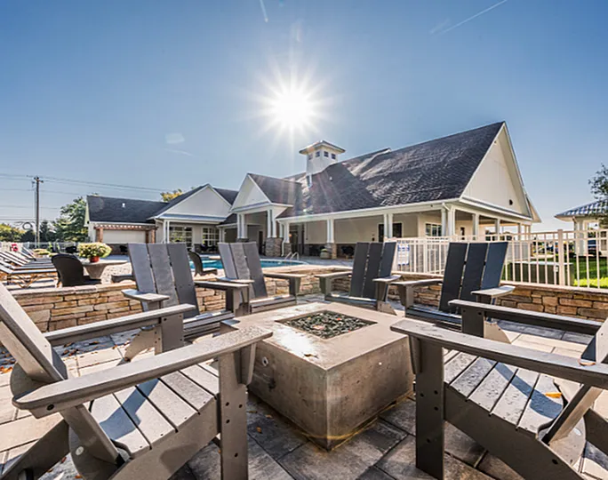 Inviting outdoor space with a cozy fire pit, stylish chairs, and a scenic view of a modern clubhouse under a bright, sunny sky.