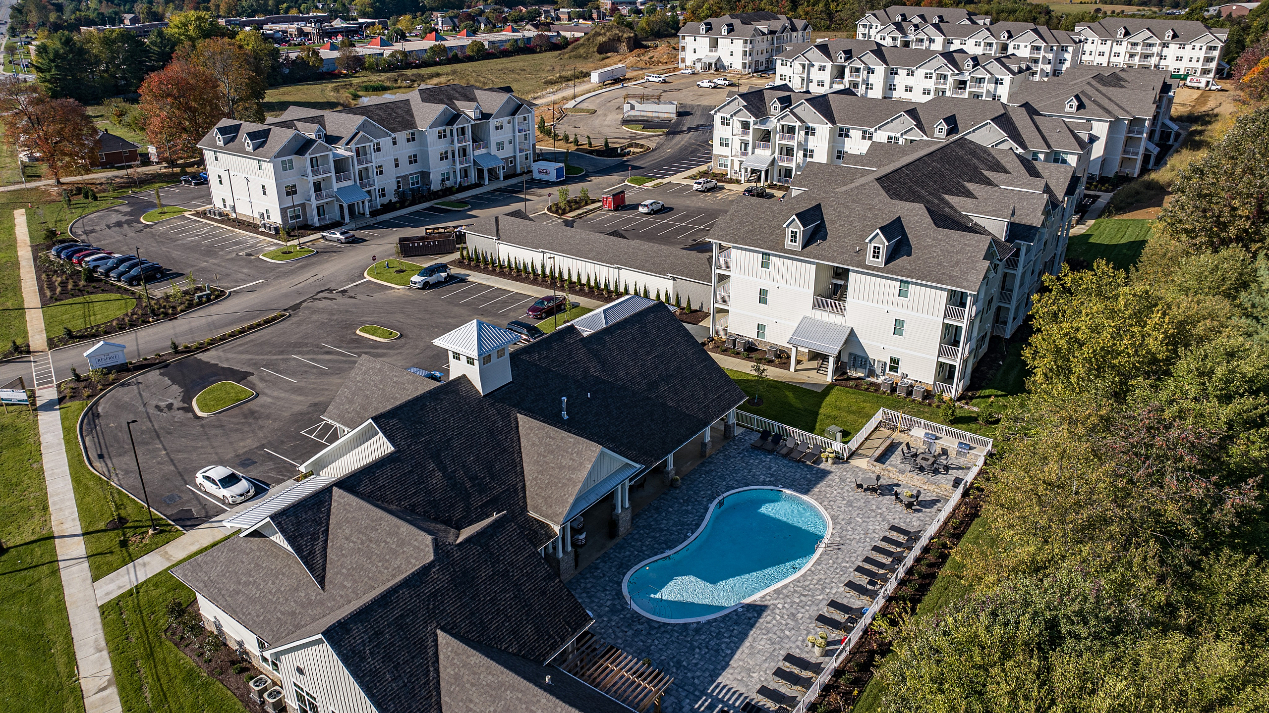 Aerial view of a modern apartment complex with spacious buildings, ample parking, and a luxurious swimming pool area surrounded by greenery.