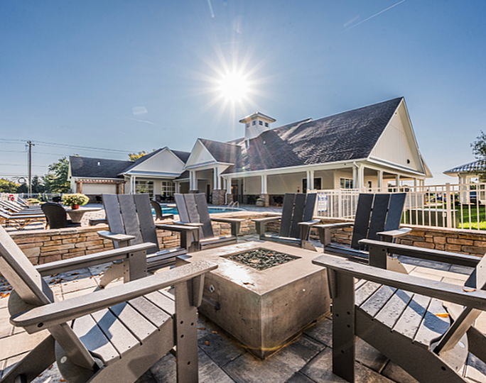 Inviting outdoor space with a cozy fire pit, stylish chairs, and a scenic view of a modern clubhouse under a bright, sunny sky.