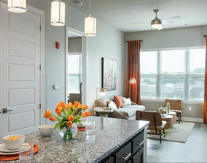 Bright and inviting apartment interior featuring a stylish granite kitchen island with vibrant tulips, leading to a cozy living area filled with natural light from large windows.
