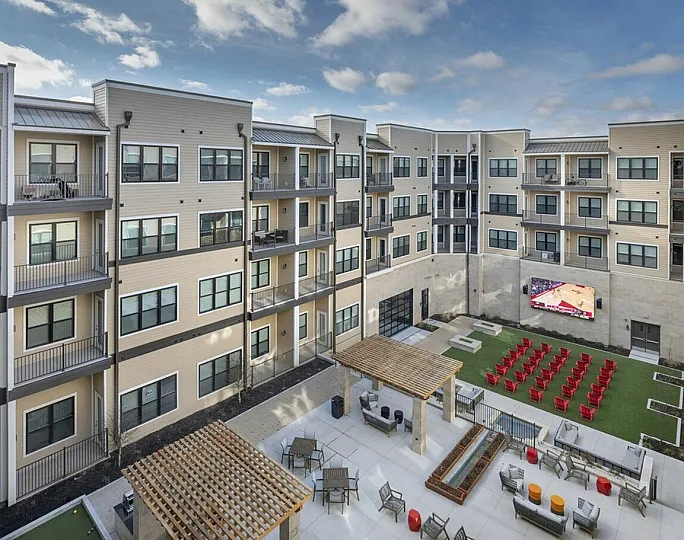 Modern apartment complex with spacious balconies overlooking a stylish courtyard featuring seating areas, a large outdoor screen, and vibrant social spaces.