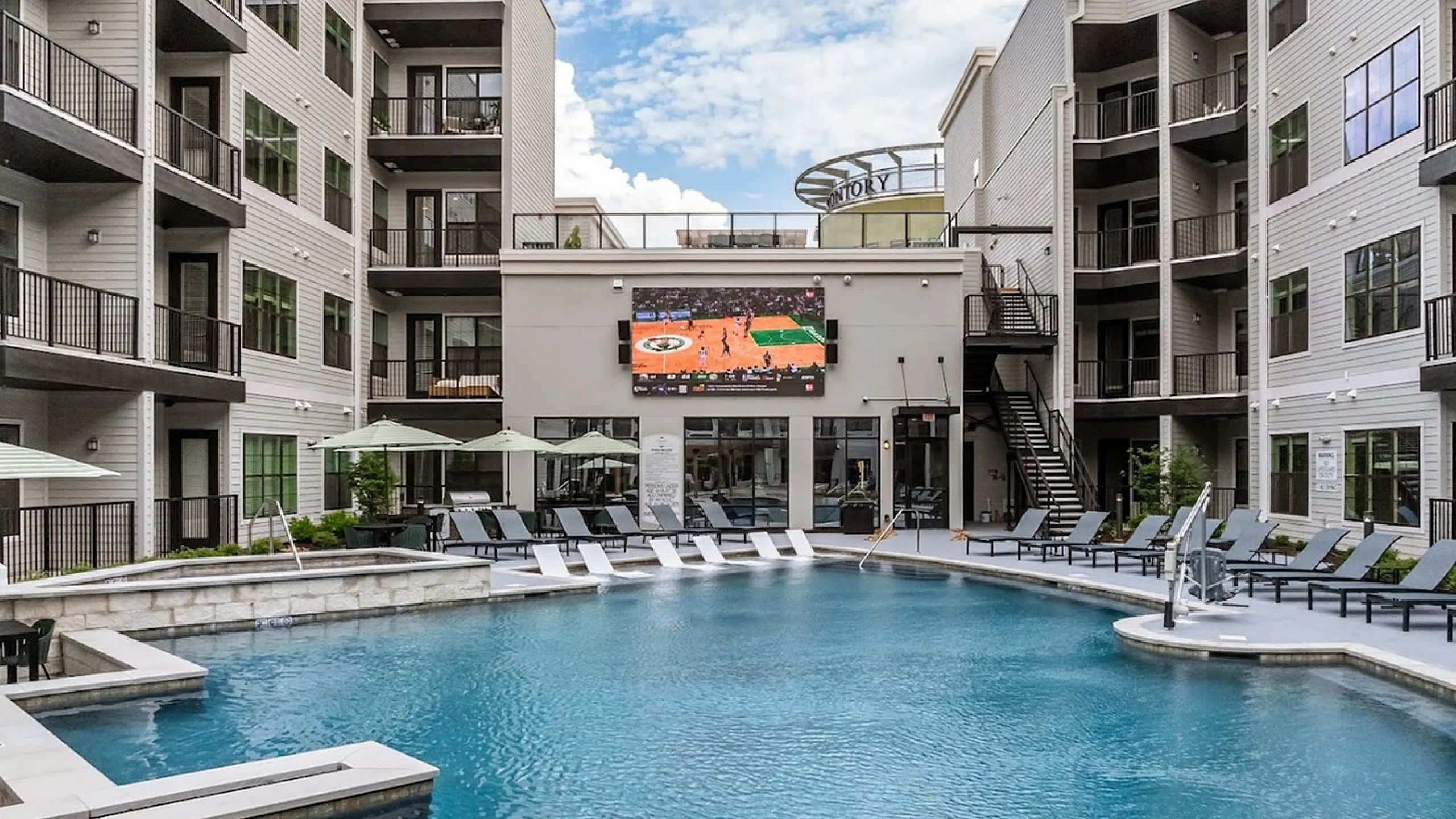 Elegant apartment complex courtyard featuring a stunning swimming pool, multiple lounge chairs, and a large outdoor screen for entertainment under the open sky.