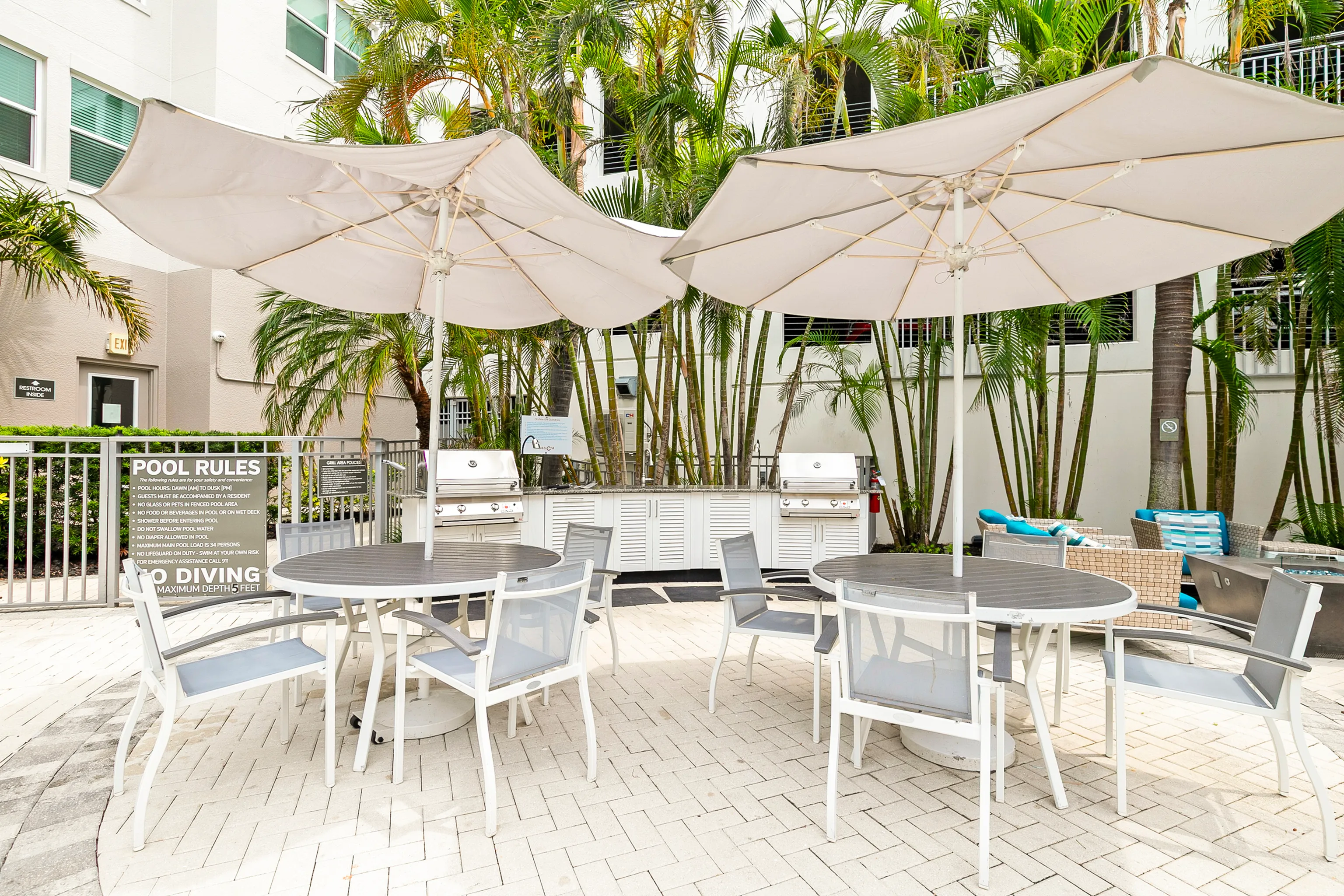 Chic outdoor patio with stylish umbrella tables, modern seating, and inviting BBQ grills surrounded by lush palm trees, perfect for social gatherings.