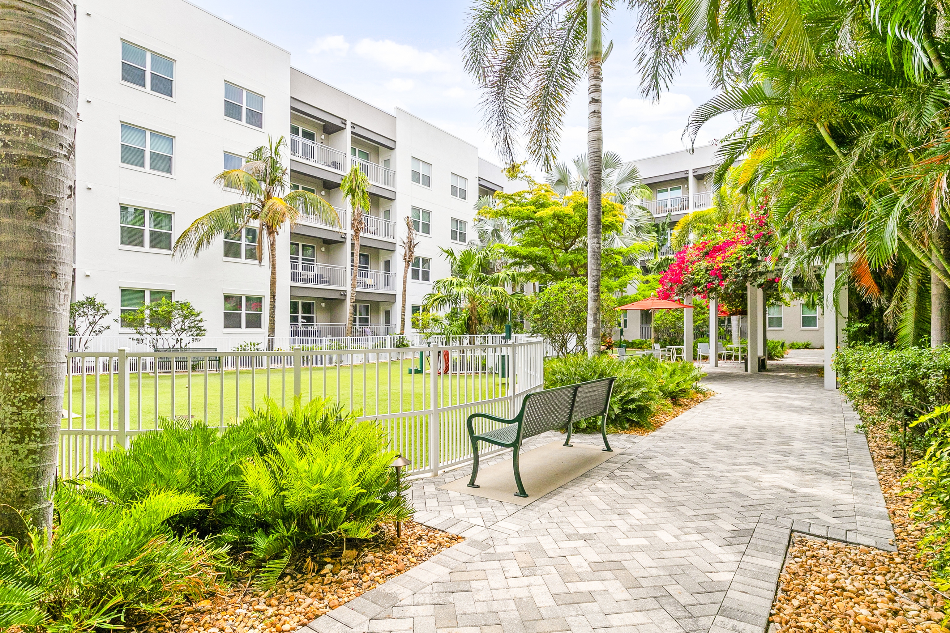 Charming apartment complex with lush landscaping, palm trees, and a peaceful walkway, offering a serene outdoor space for residents.