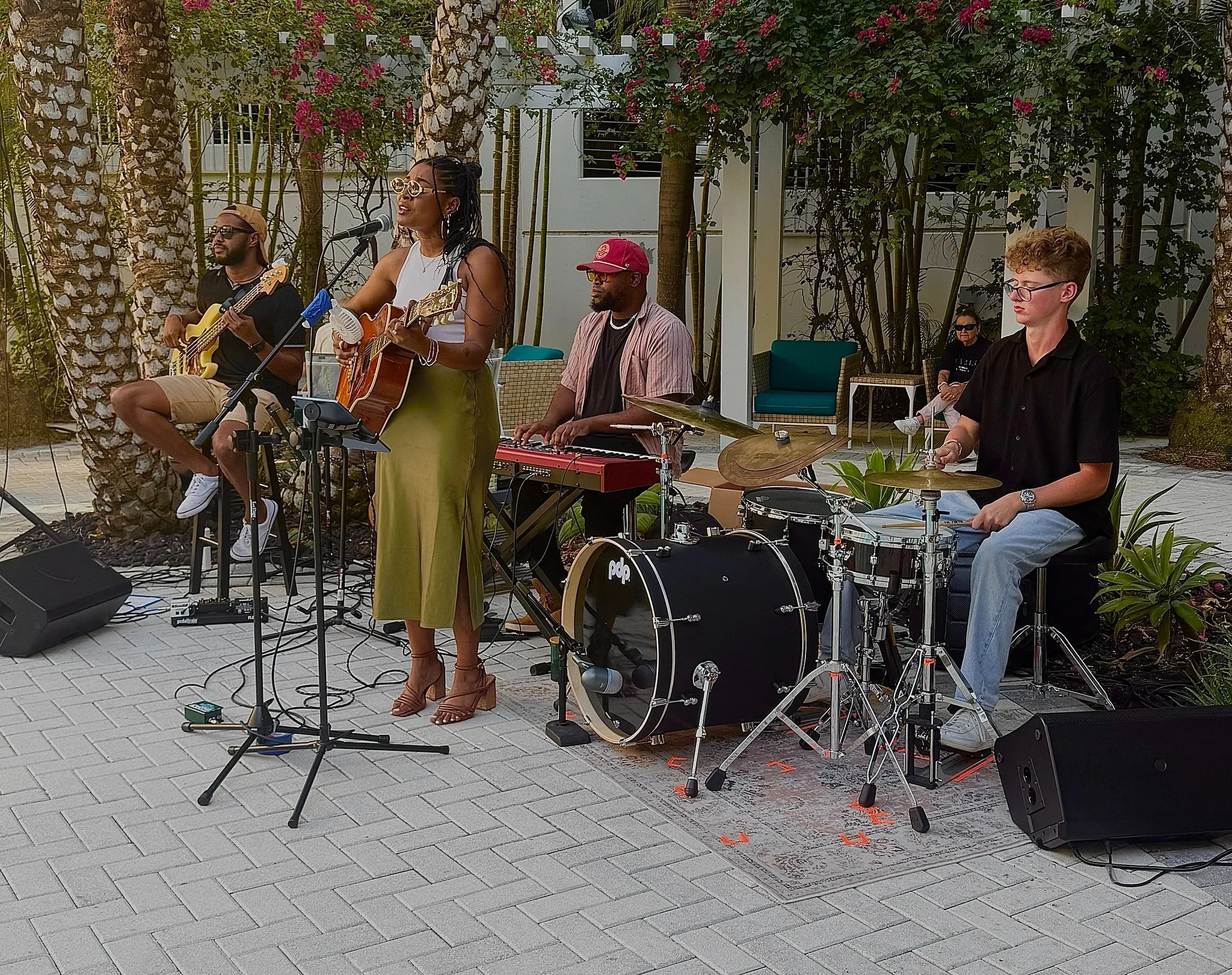 Vibrant outdoor band performance with a singer-guitarist, keyboardist, bassist, and drummer under lush palm trees.