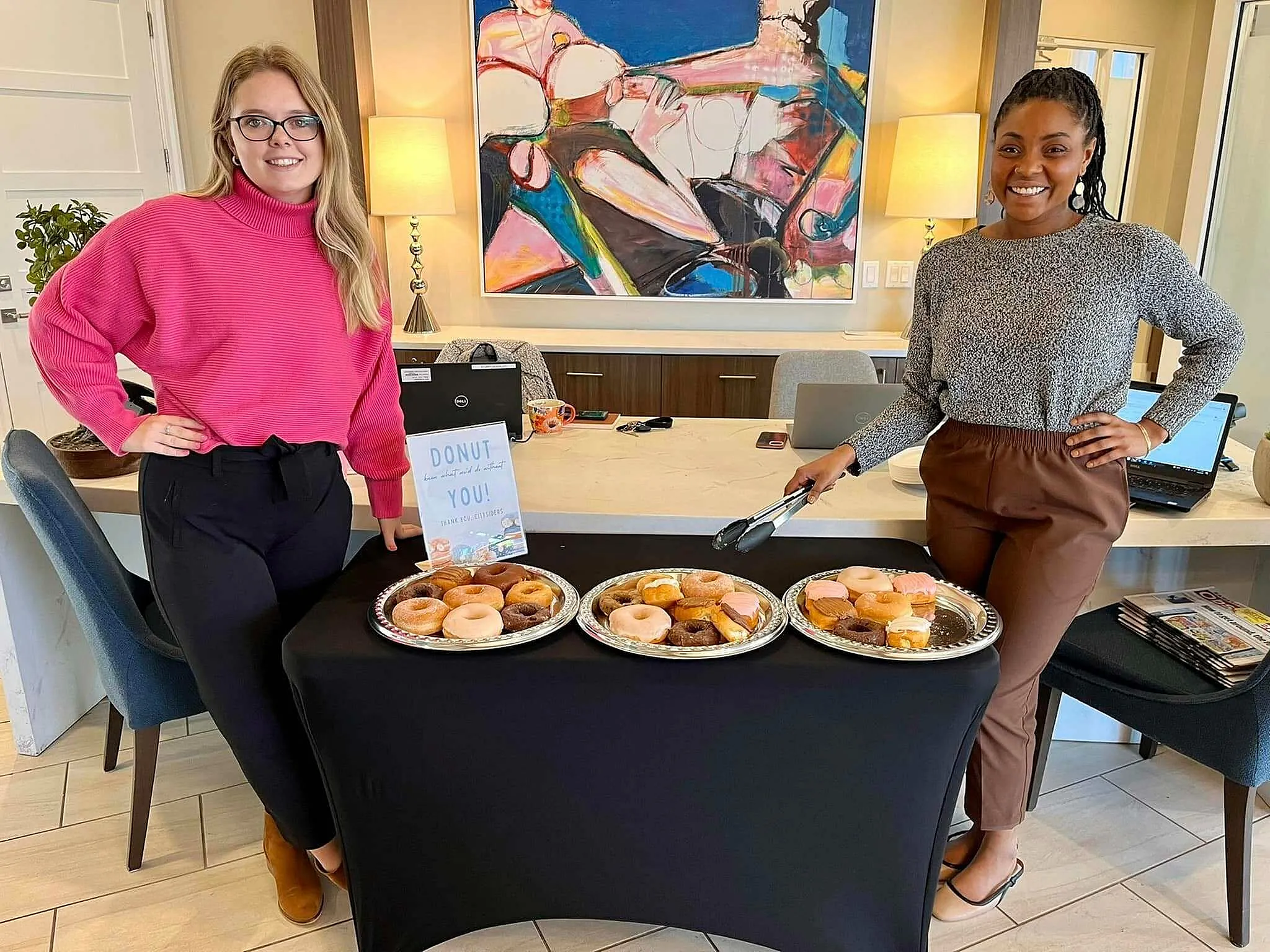 Inviting apartment welcome event featuring two smiling hosts with trays of colorful donuts in a modern, stylish lobby setting.