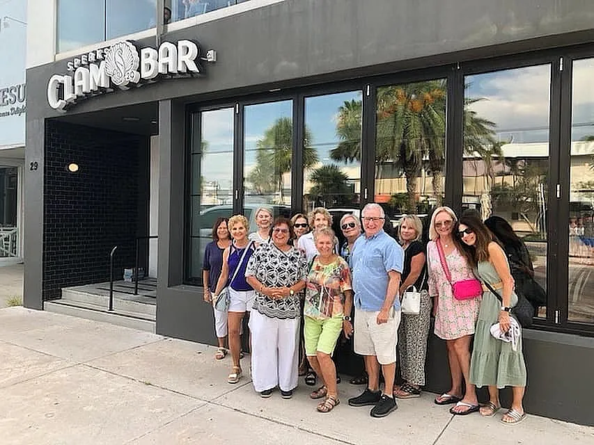A group of cheerful people posing in front of a modern clam bar with large windows, reflecting a sunny day with palm trees in the background.