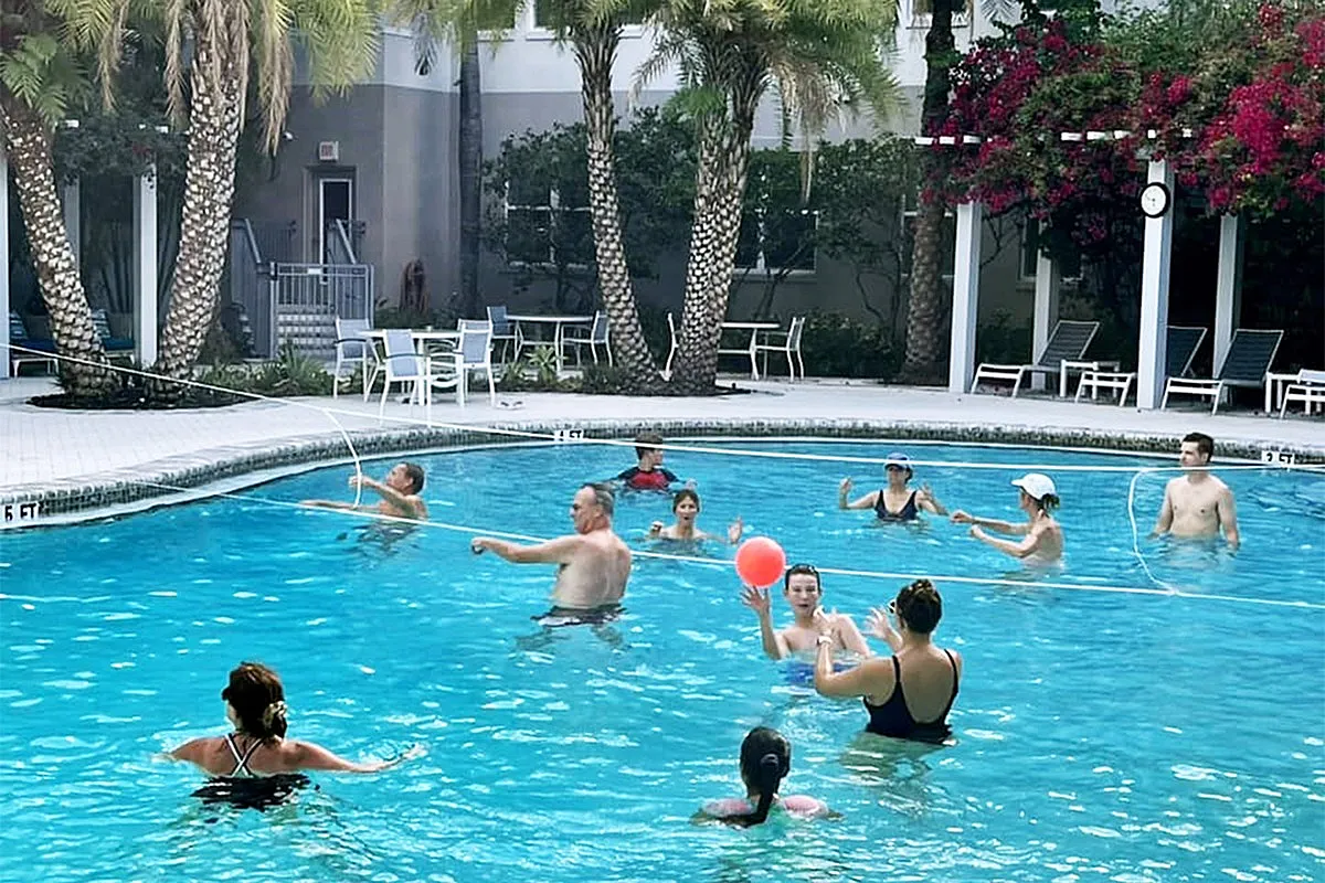 Vibrant community pool scene with people enjoying a game in crystal-clear waters, surrounded by lush palm trees and elegant loungers.