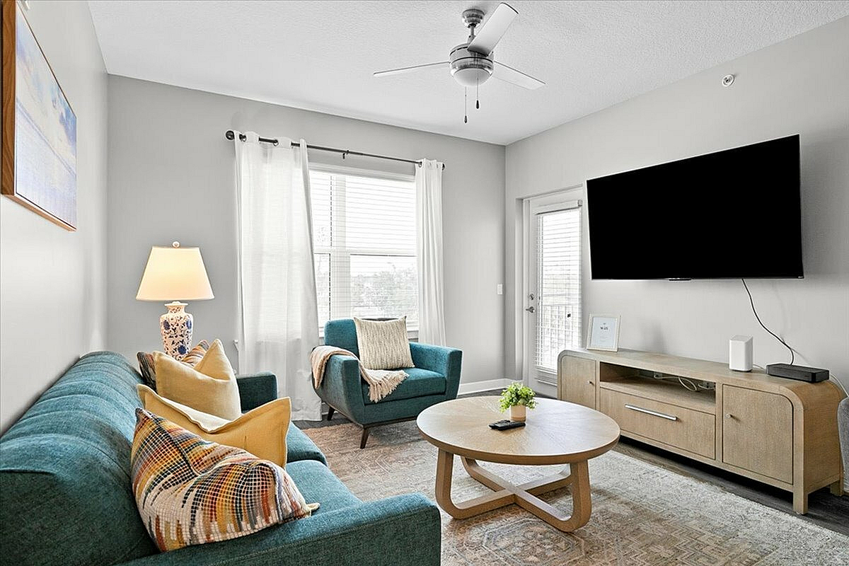 Bright and cozy living room featuring a teal couch and armchair, chic round coffee table, elegant lamp, large window, and modern TV setup. | City Side Apartments | Sarasota, FL
